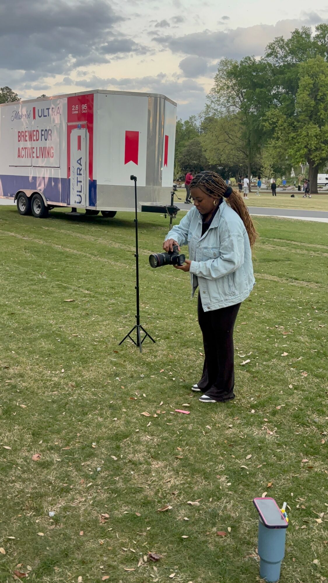 Woman with curly hair in a light jacket holding a camera outdoors on grass, with a truck and trees in background.