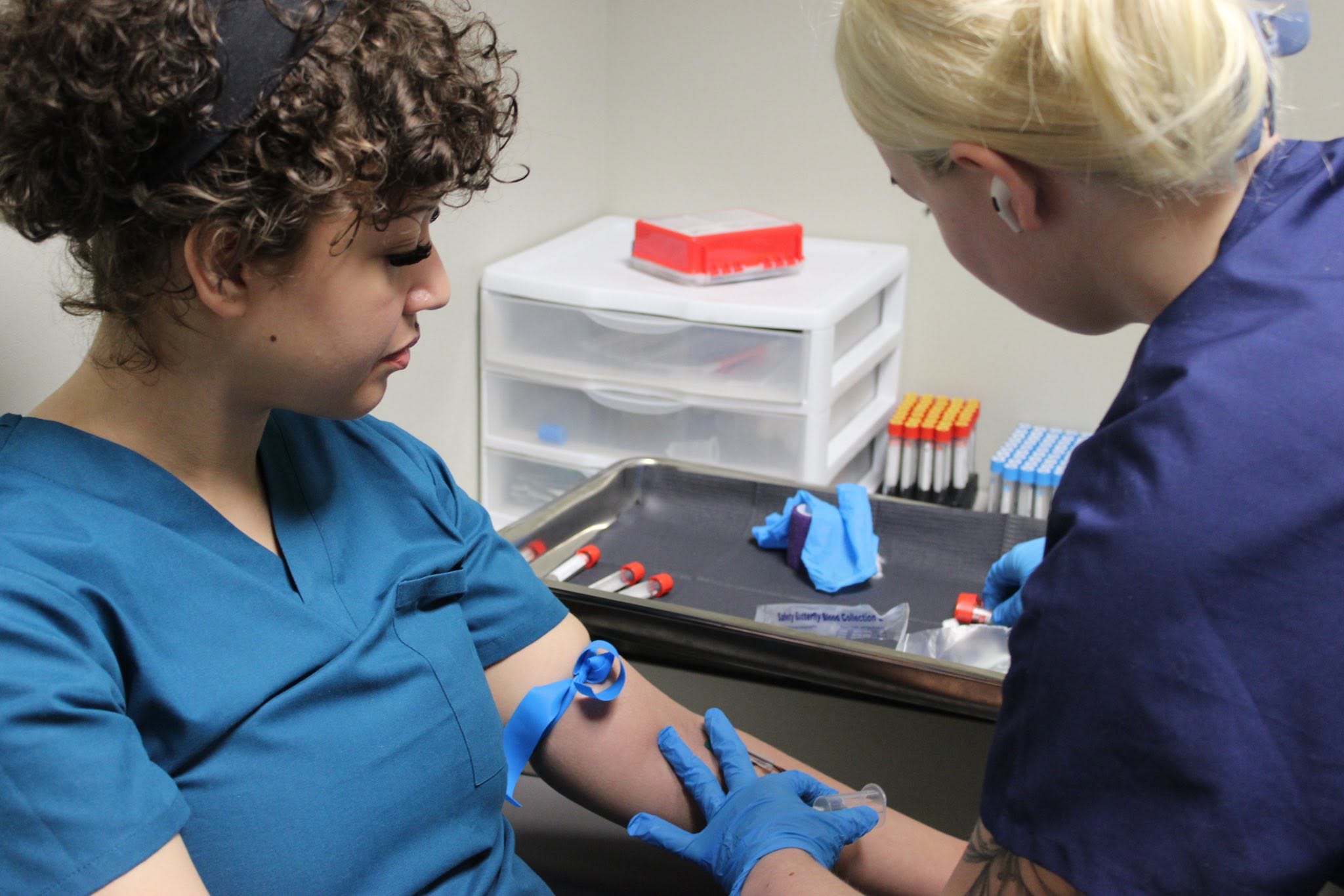 Two healthcare workers in blue scrubs and gloves preparing a patient's arm for a medical procedure in a clinical setting.