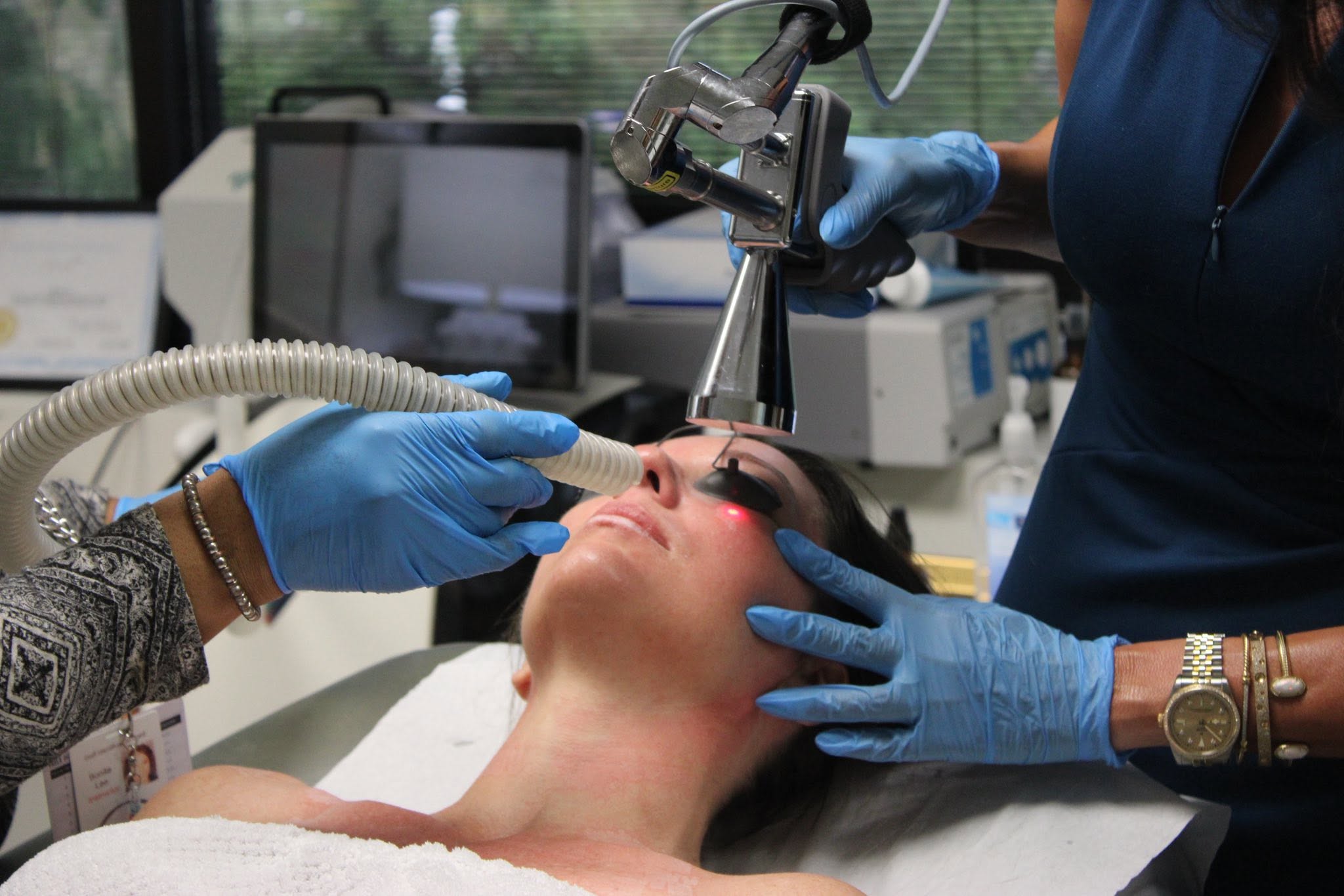 Medical professional performs a procedure on a woman lying down, with equipment and another person assisting, in a clinical setting.