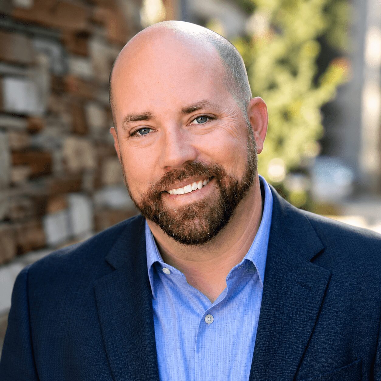 Man with a beard and bald head smiling outdoors, wearing a blue suit jacket and light blue shirt, with a blurred background.