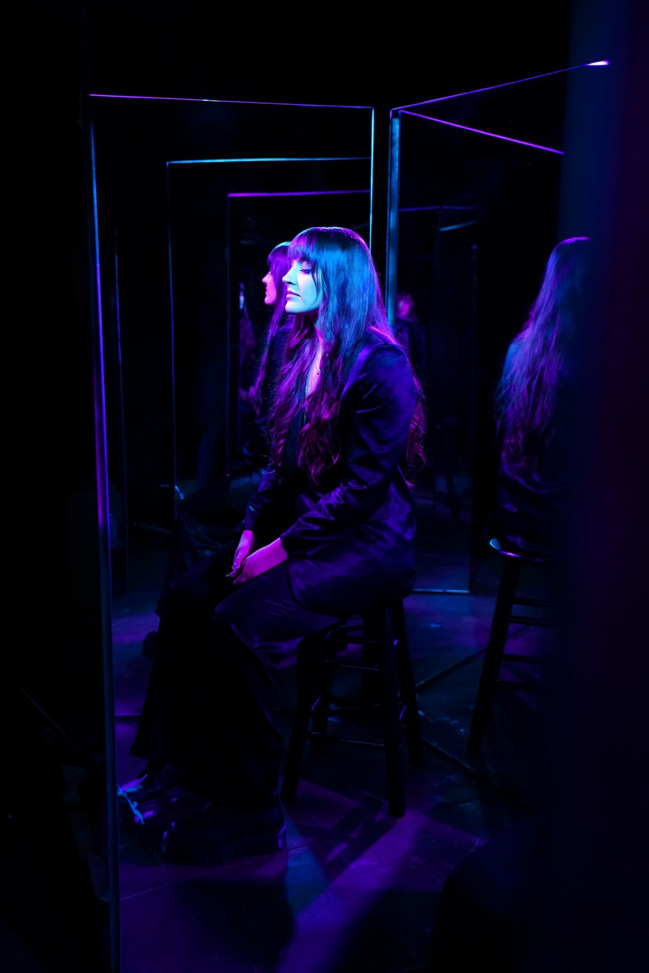 Young woman with long hair sitting in a dark room illuminated by purple and blue neon lights, surrounded by mirrors.