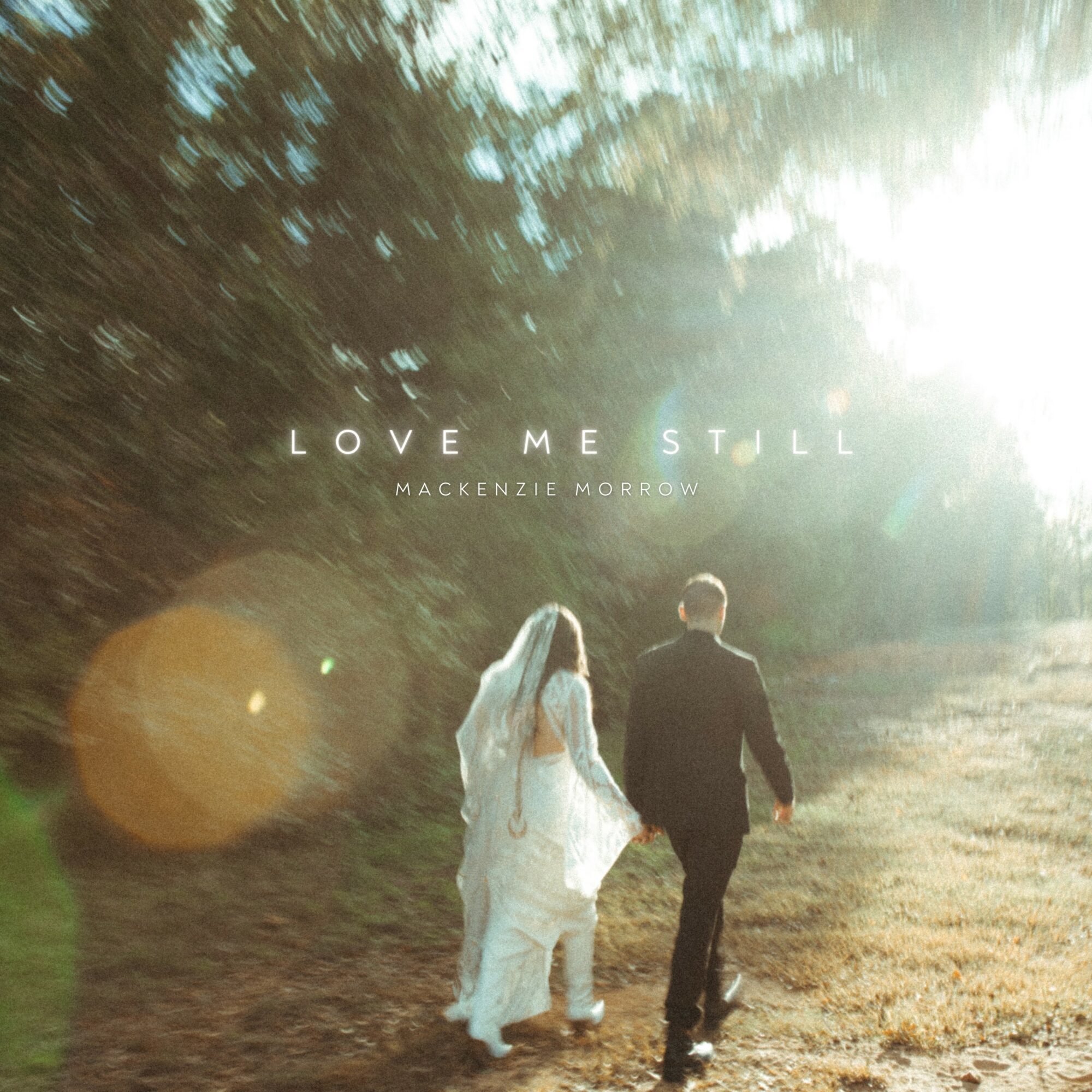 A couple walking outdoors on a sunny day, with trees and sunlight in the background, holding hands. Text reads 'Love Me Still' and 'Mackenzie Morrow'.