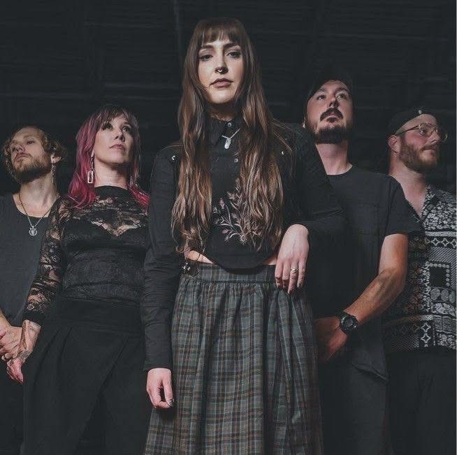 Group of five people standing together, dark background, woman in front with long hair and plaid skirt, others behind her.