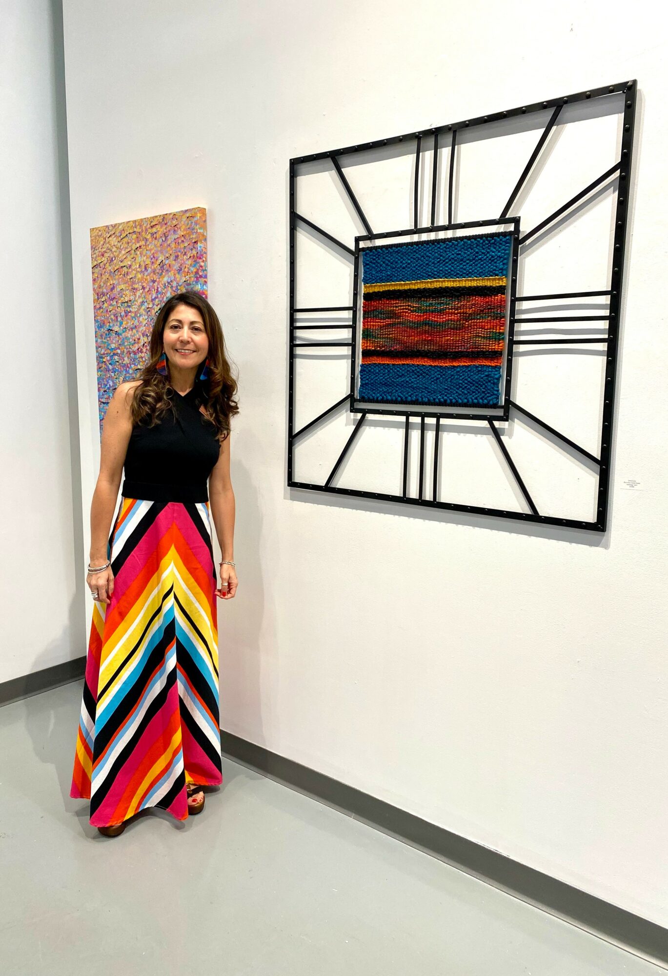 Woman in colorful striped dress standing next to a large wall clock with a colorful artwork in the center, in an art gallery.
