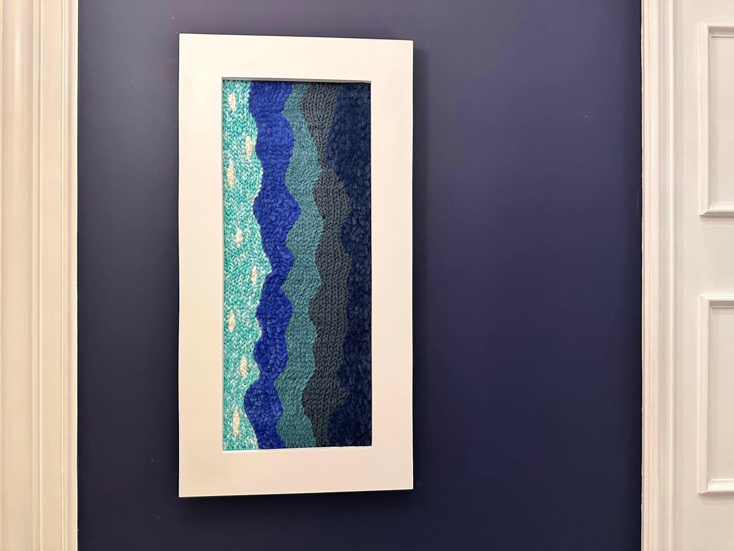 Vertical framed abstract artwork with wavy blue, teal, and white patterns on a dark purple wall.