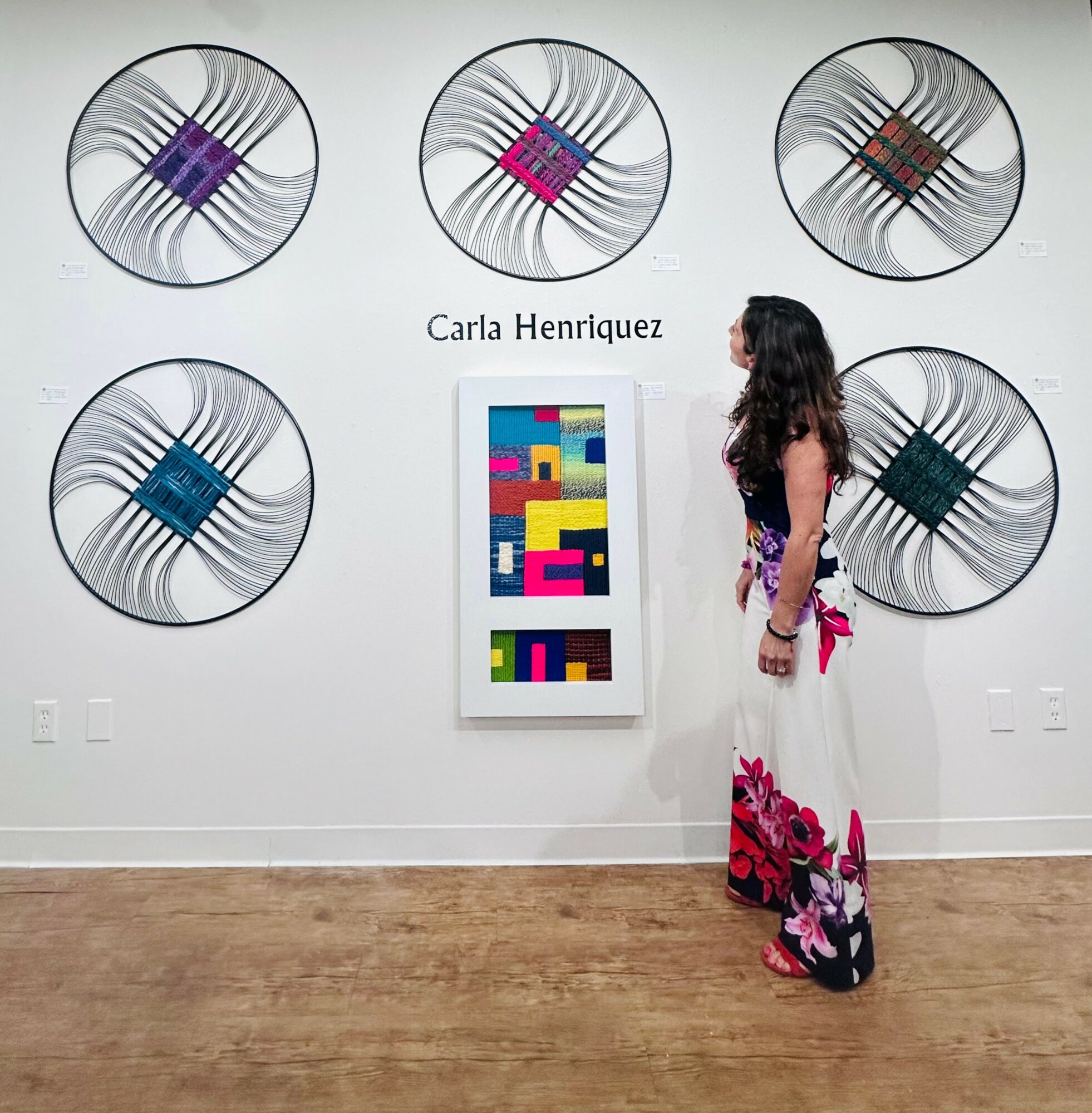 Woman in a floral dress looking at colorful artwork on a white wall with six circular art pieces above.