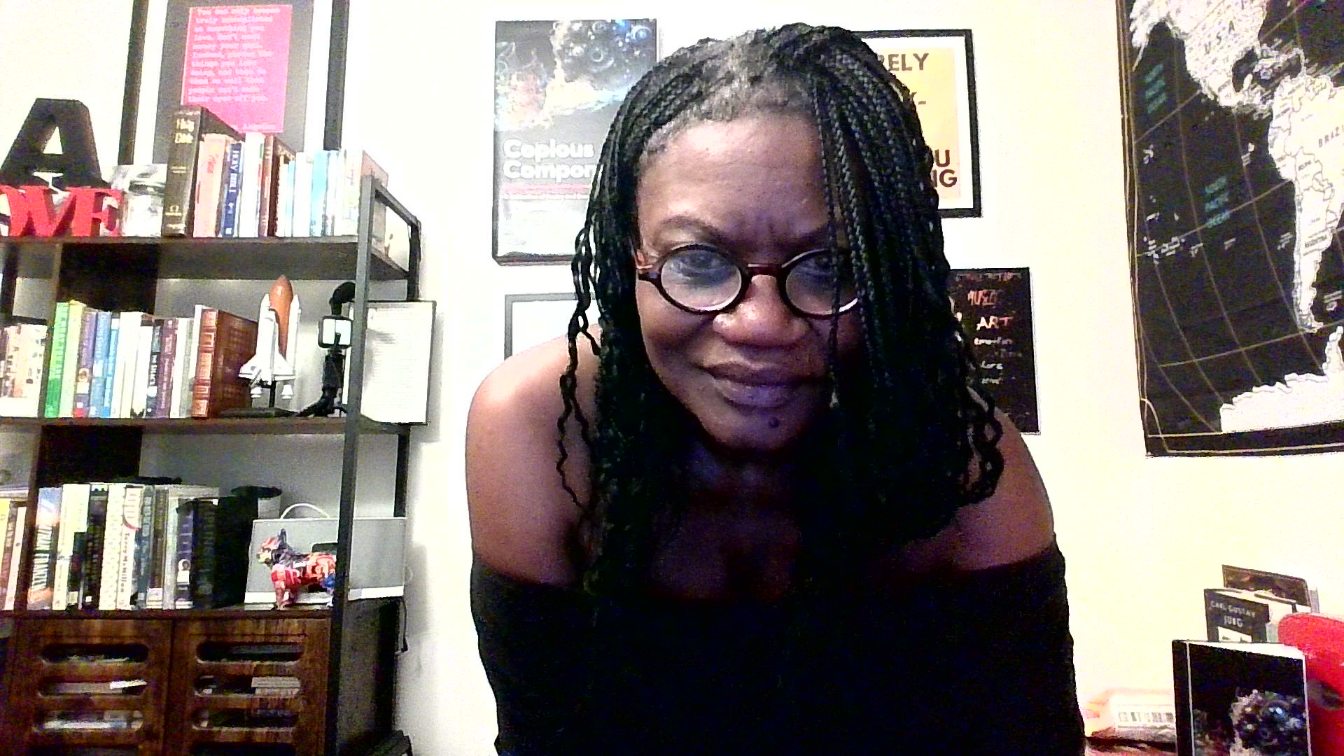 Woman with glasses and braided hair smiling indoors, bookshelf and framed pictures in background.