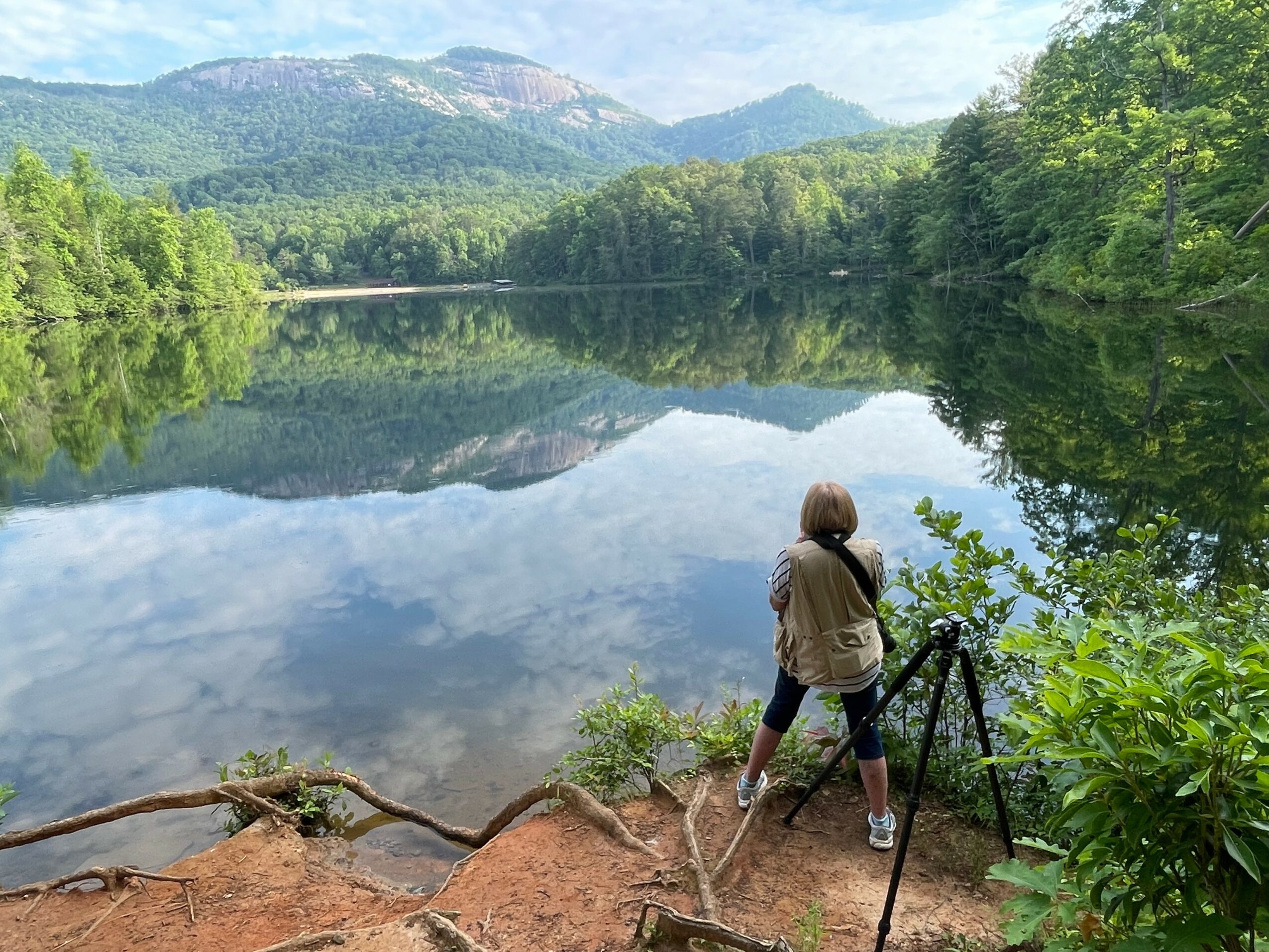 Person standing on riverbank with camera on tripod, overlooking calm lake surrounded by green trees and mountains.