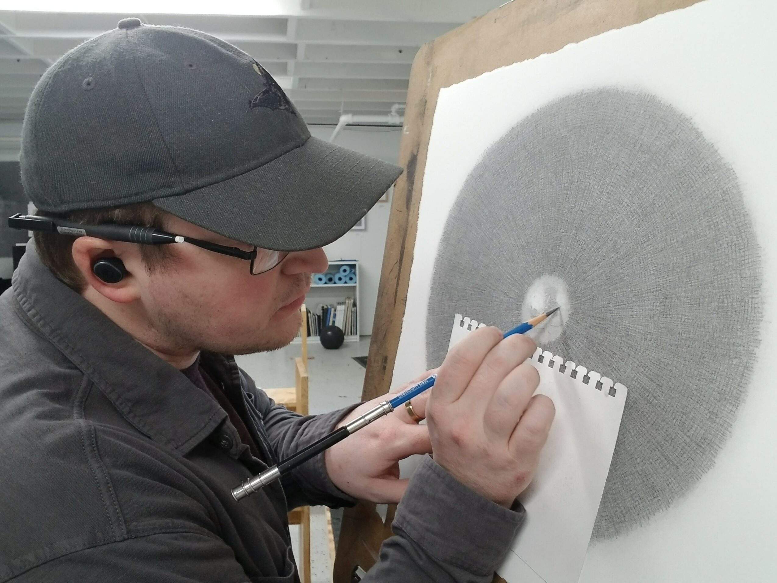 Person wearing glasses and a cap painting a large gray circle on paper attached to a wooden board.