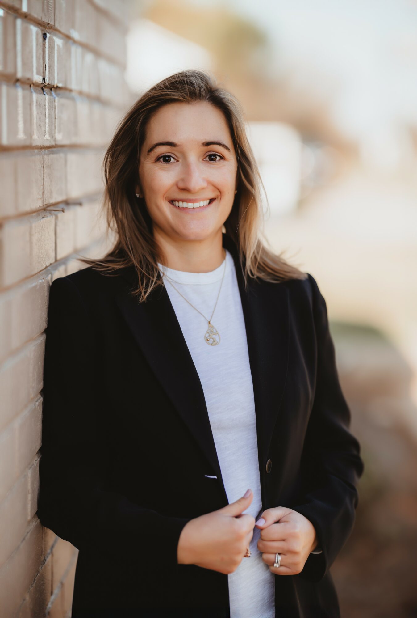 Young woman with shoulder-length hair smiling, standing outdoors next to a brick wall, wearing a black blazer and white shirt.