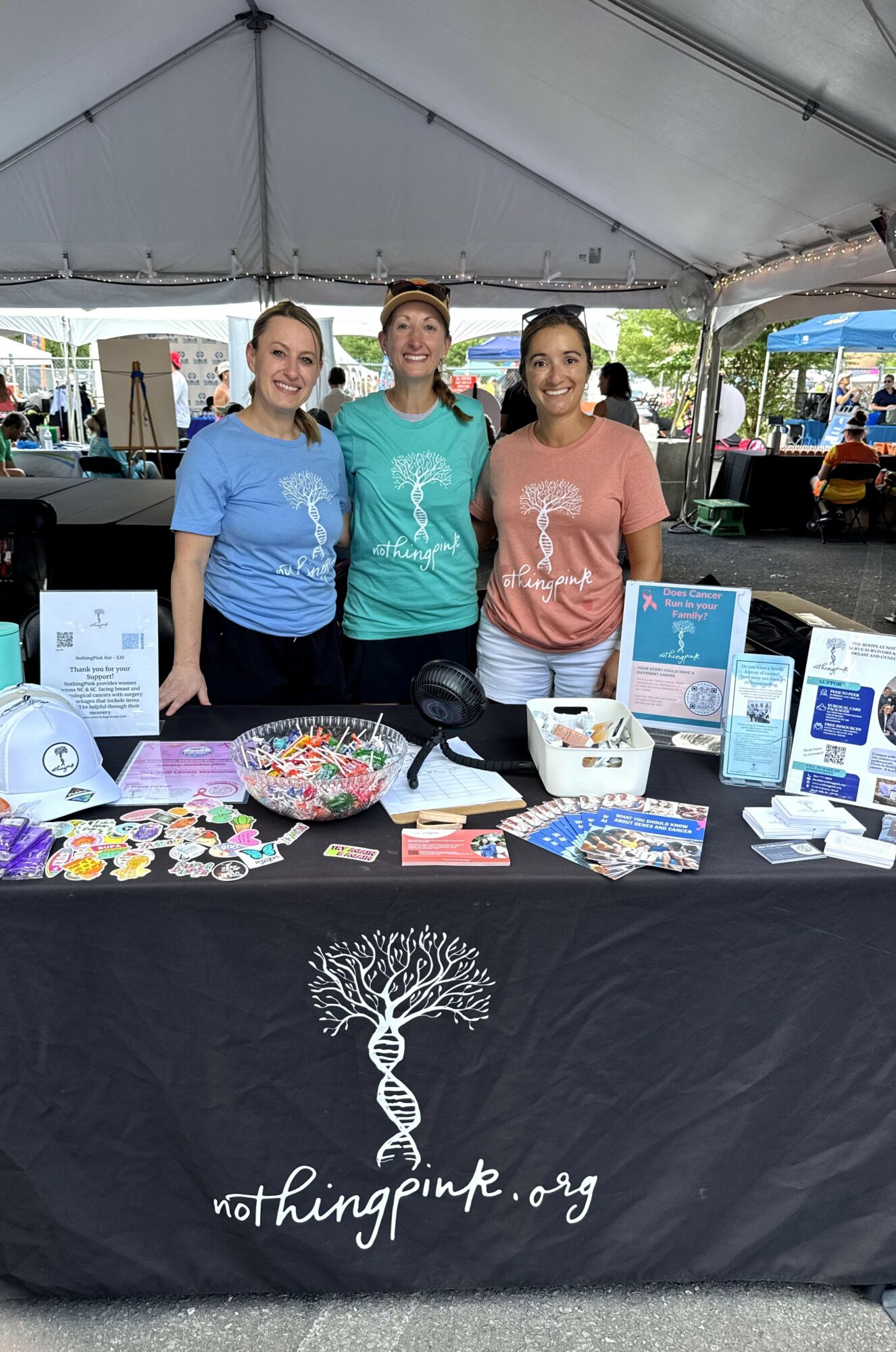 Three women stand behind a table with informational materials and a banner for nothingpink.org under a tent.