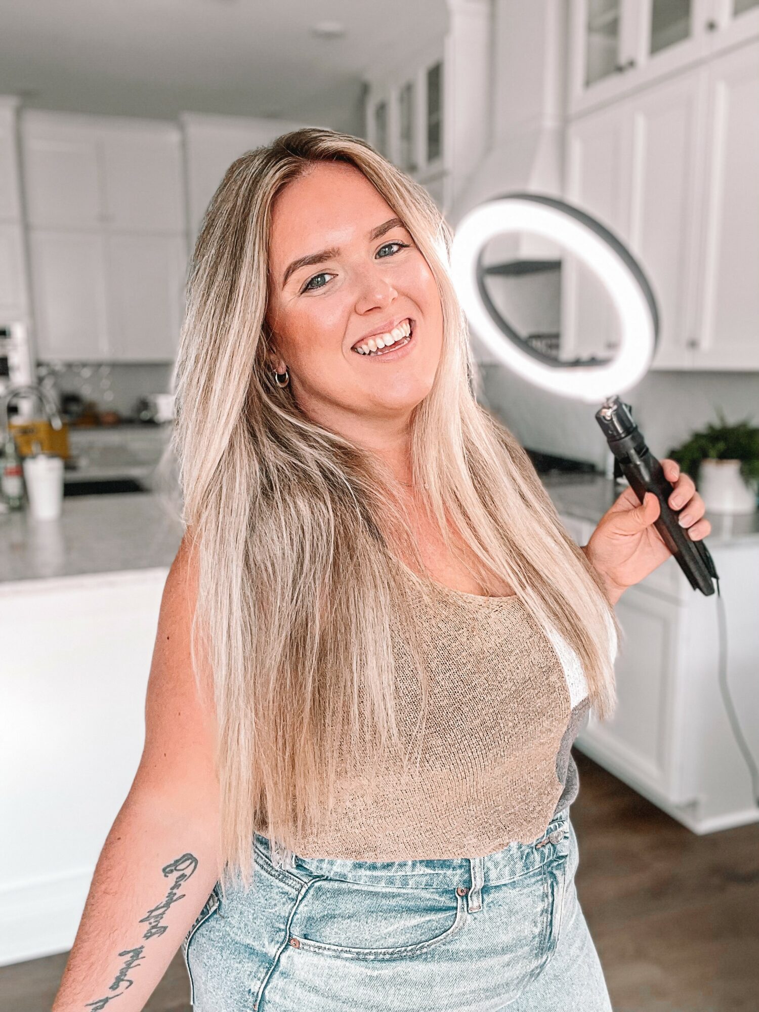 Woman with long blonde hair smiling, holding a ring light in a bright kitchen, wearing a beige top and jeans.