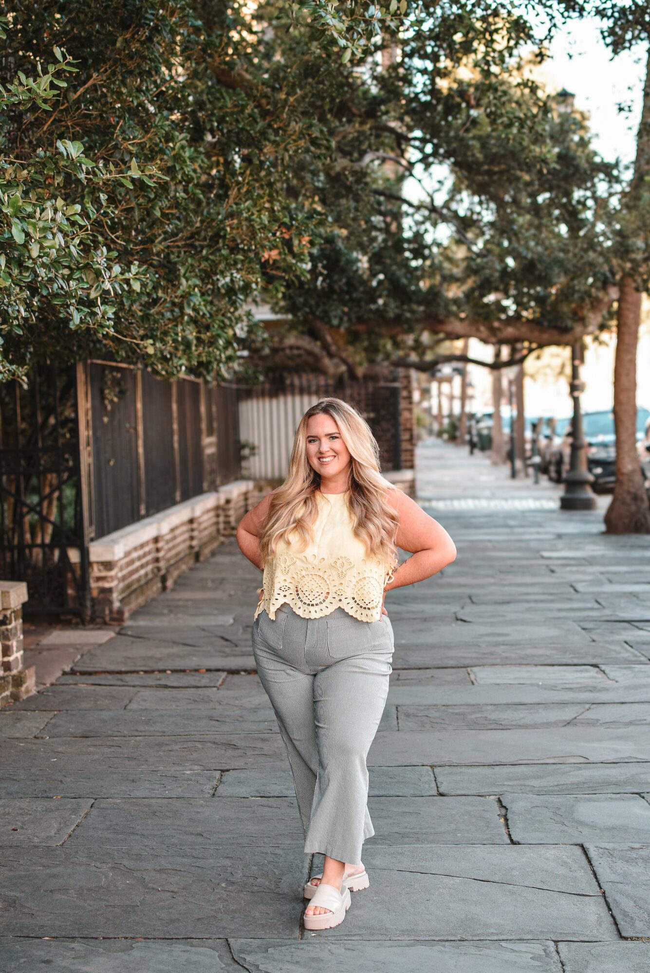 Woman walking on sidewalk with trees and buildings in background, smiling, wearing a yellow top and gray pants.