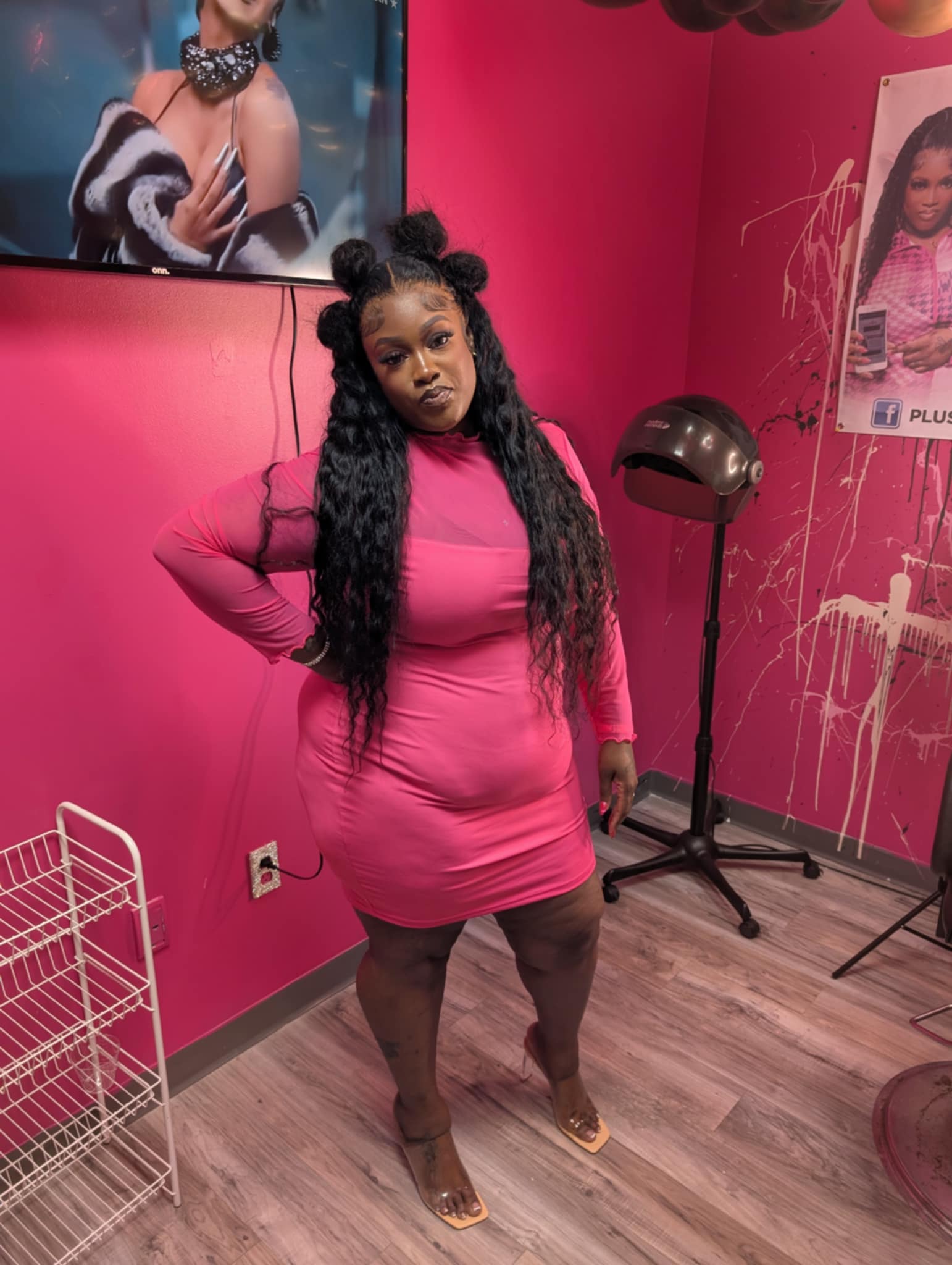 Woman in pink dress with long curly hair standing in salon with pink walls and hair styling tools.
