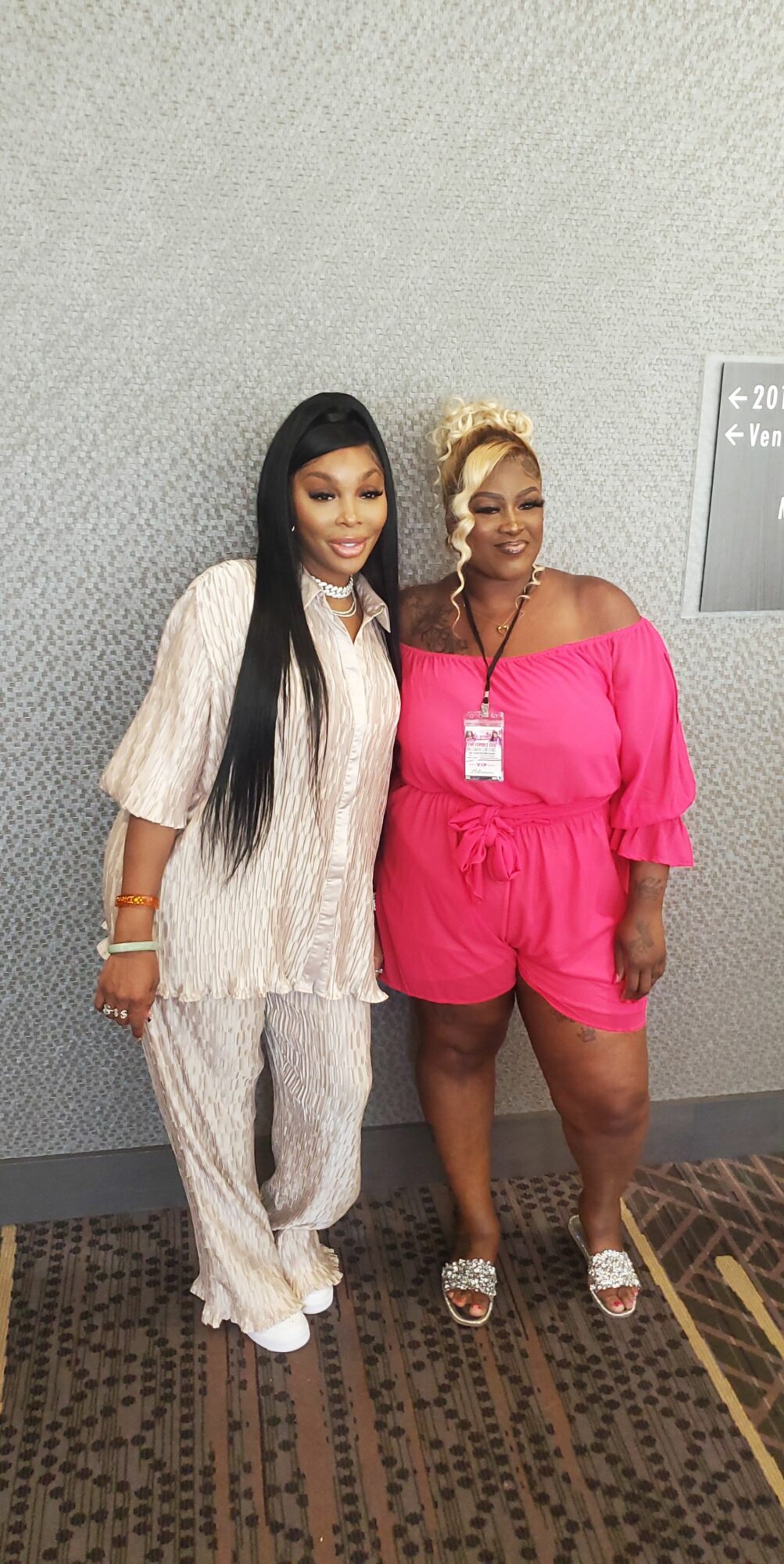 Two women standing side by side against a gray wall, one in a white outfit, the other in a pink dress, smiling.