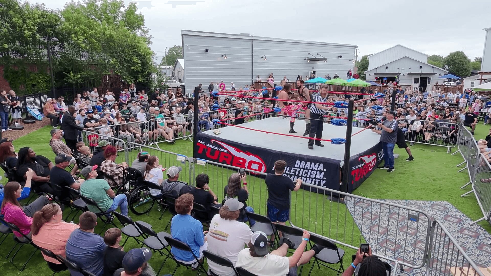 Professional wrestling match outdoors with audience seated around a ring, surrounded by metal barriers, on a grassy field.
