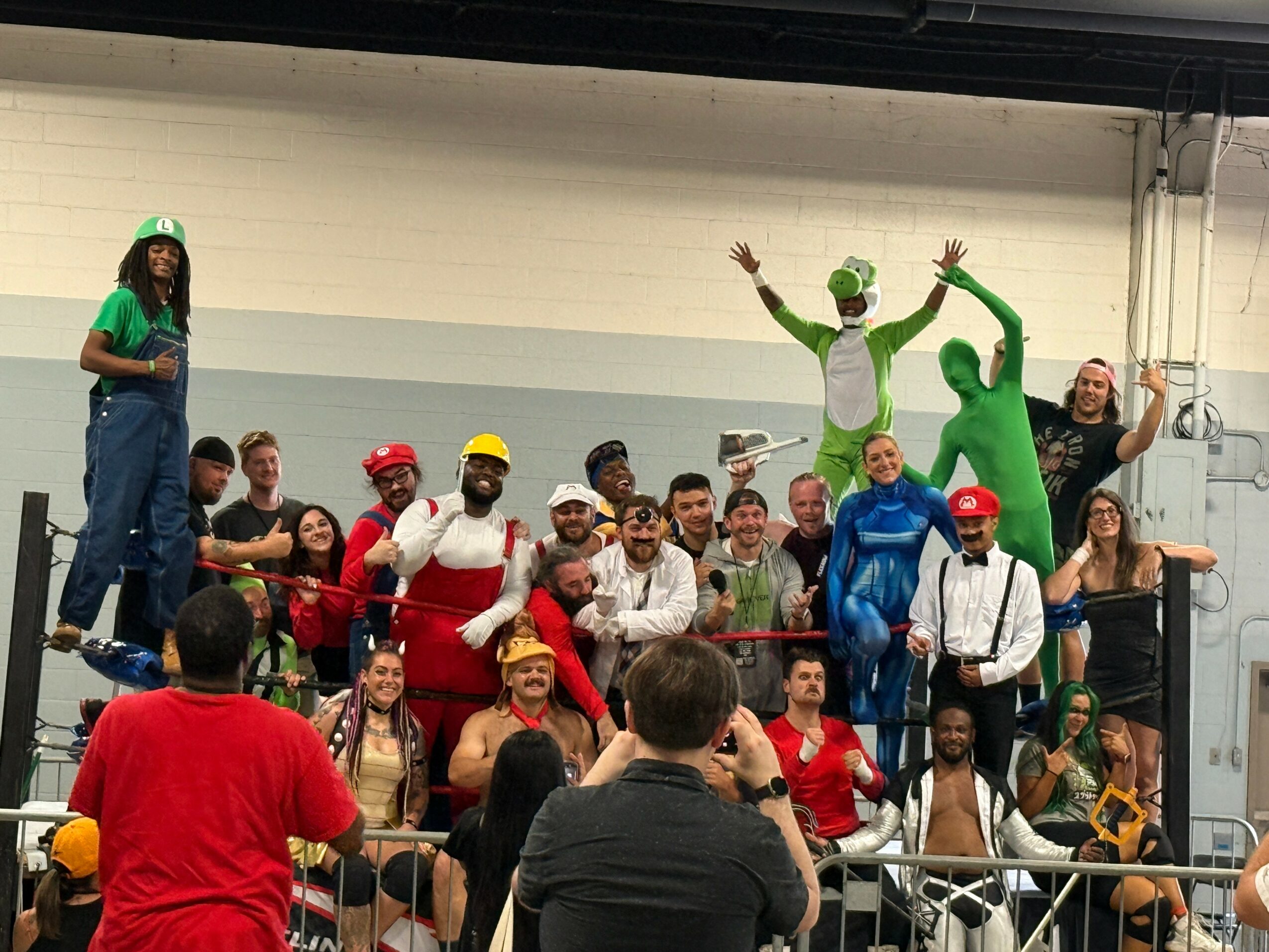 Group of people in colorful costumes and casual clothing posing indoors, some standing on a platform, others sitting or kneeling, with a person taking a photo.