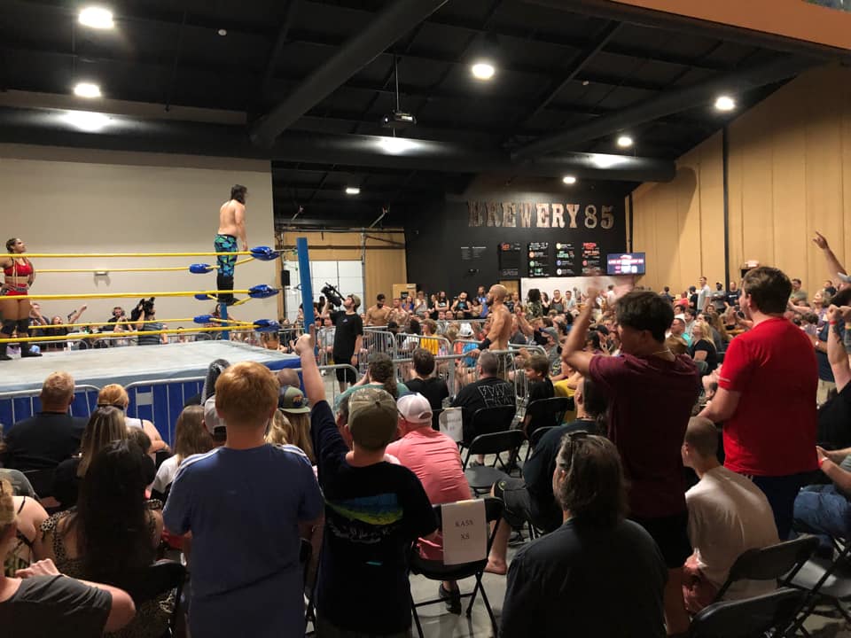 Wrestling match in a ring with audience watching, some standing and cheering, in a large indoor venue.