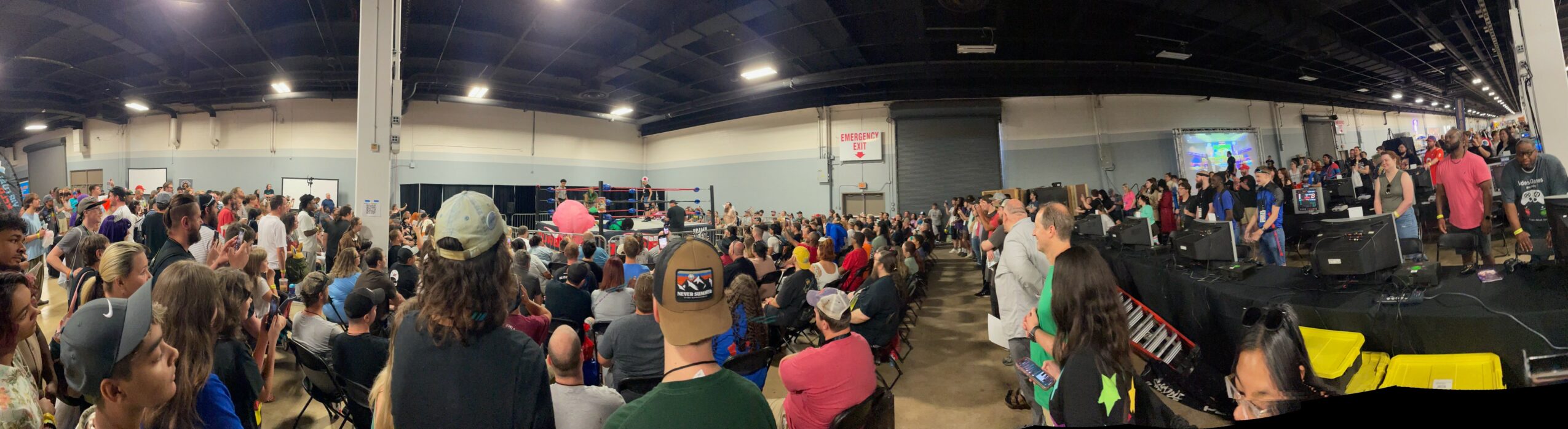 Large indoor gathering with many people seated and standing, stage with speakers and screens at the front, ceiling with lights.