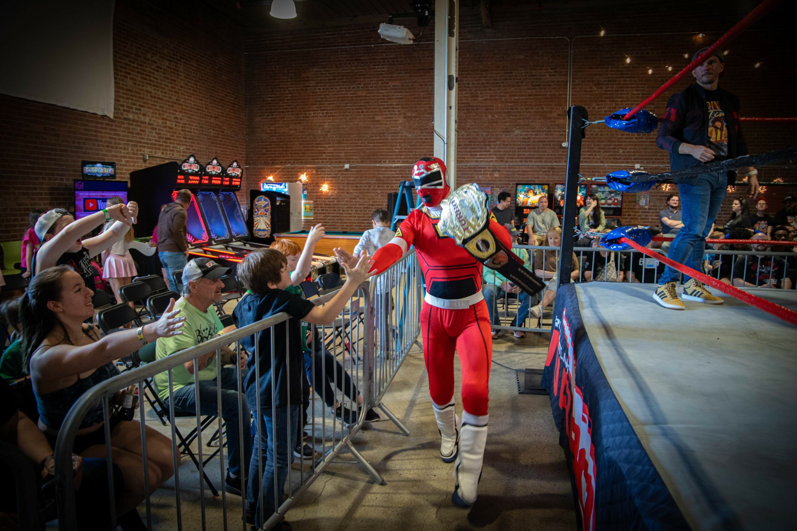 Wrestler in red and white costume high-fives children behind a barrier in a wrestling ring.