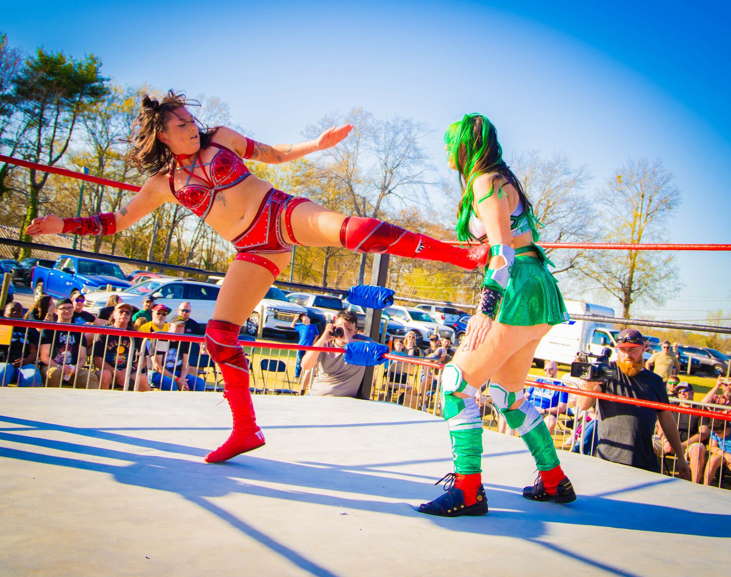 Two women wrestle in a ring outdoors with a crowd watching, one in red and the other in green costume.