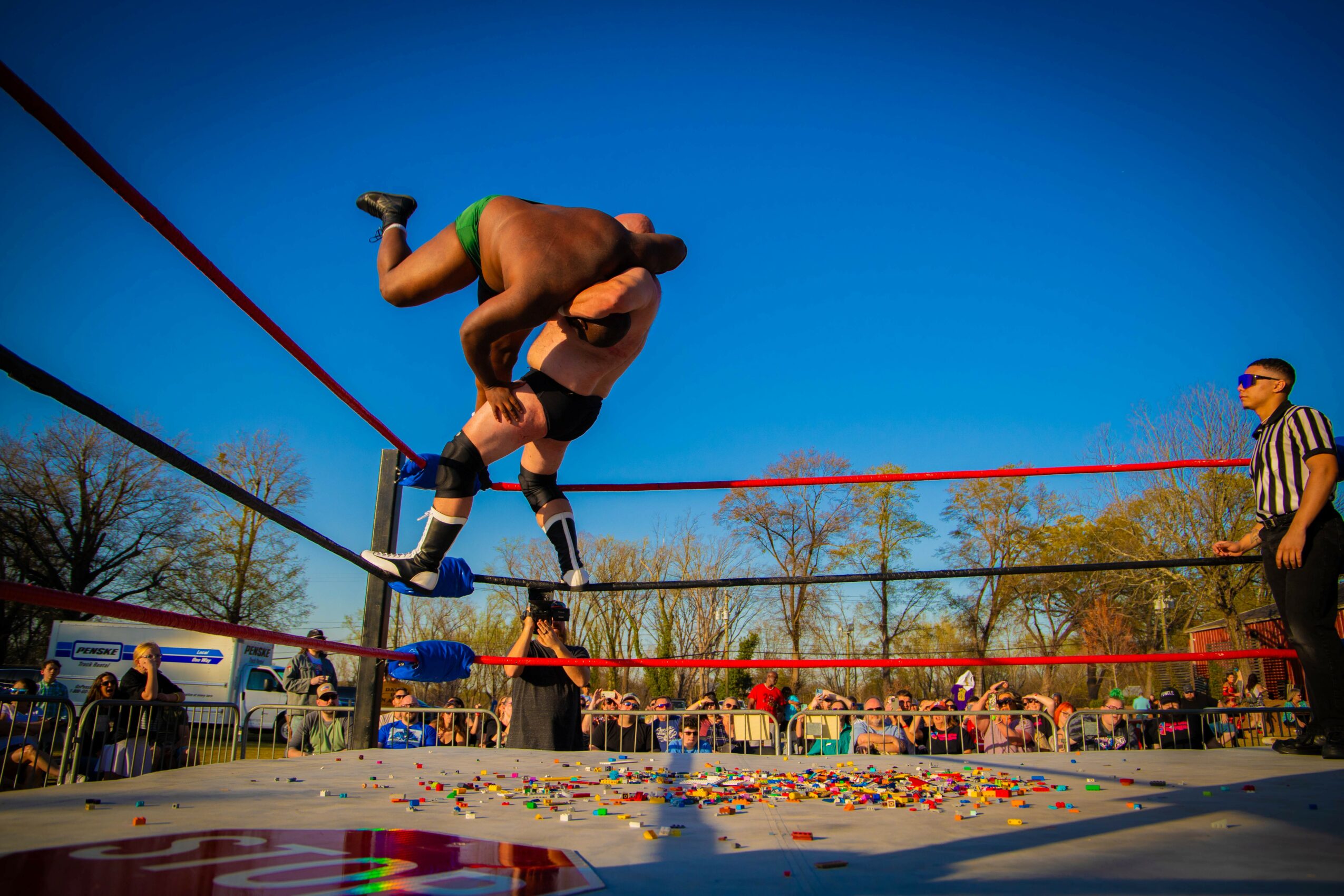 Two wrestlers in ring, one lifting other upside down, referee and audience watching outdoors, colorful confetti on ground.