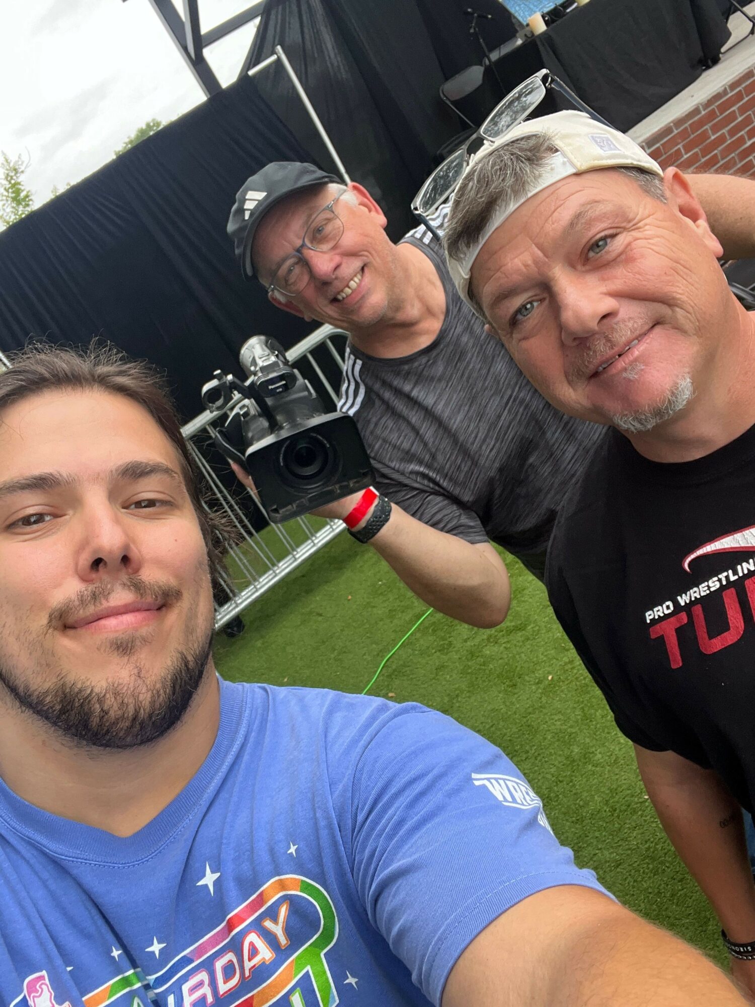 Three men outdoors, one with glasses and a cap, another with glasses, and a third with a beard, smiling.