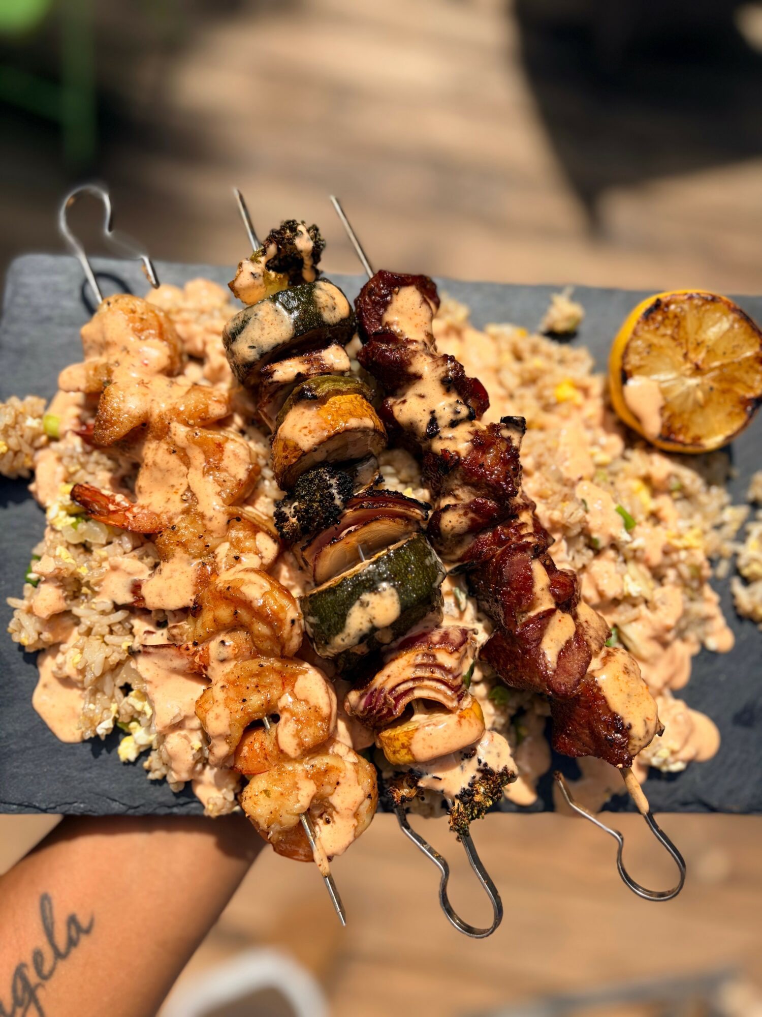 Skewers of grilled vegetables and meat on a bed of rice, with a lemon half on the side, outdoors.