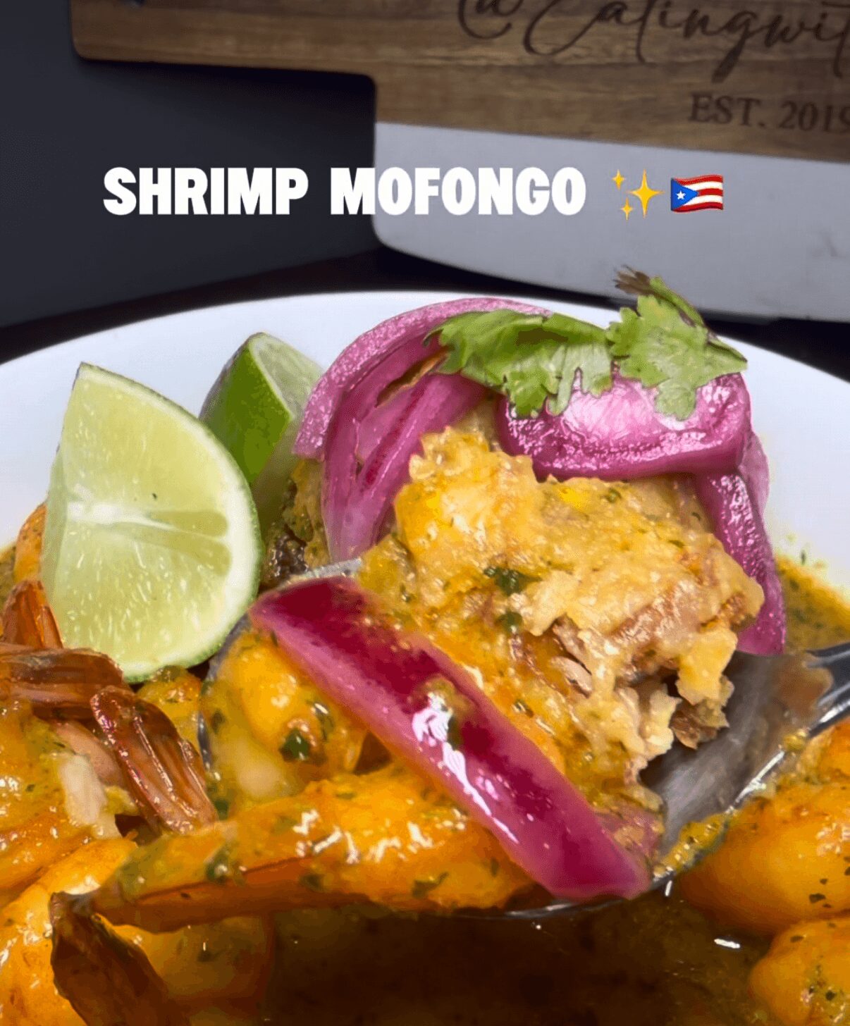 Close-up of a shrimp mofongo dish with lime wedges, purple onions, and a fork, on a dark surface.