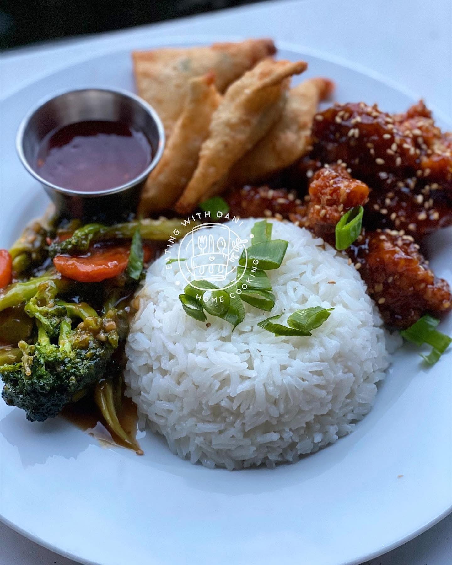 Plate with white rice, fried chicken, broccoli, carrots, and dipping sauce, garnished with green onions.