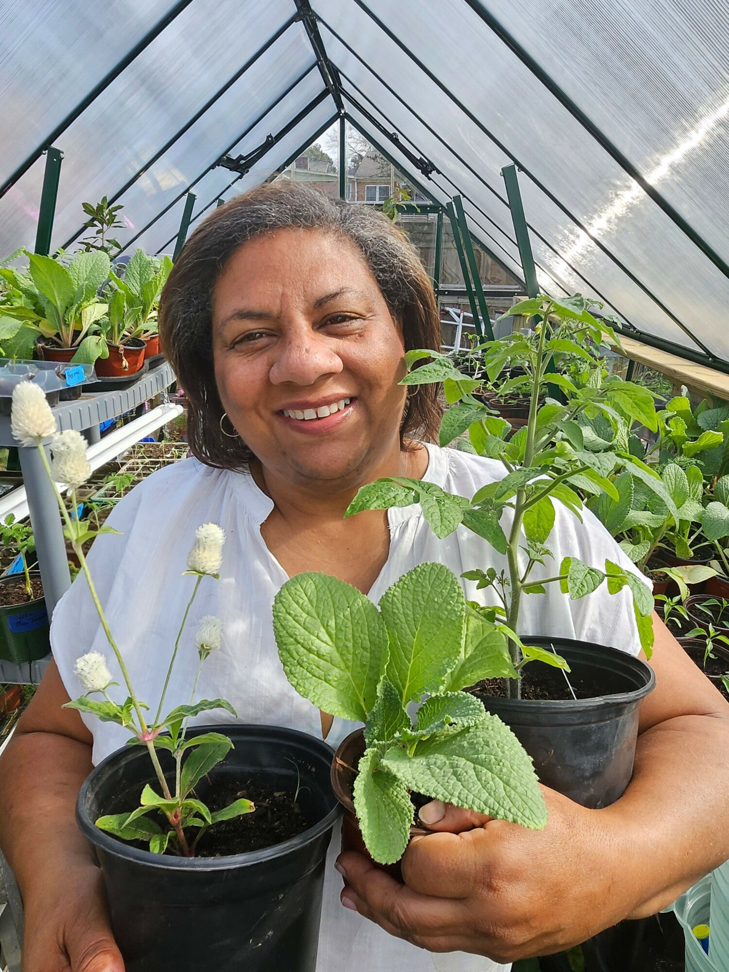 Smiling woman holding potted plants in a greenhouse, surrounded by green foliage and gardening tools.