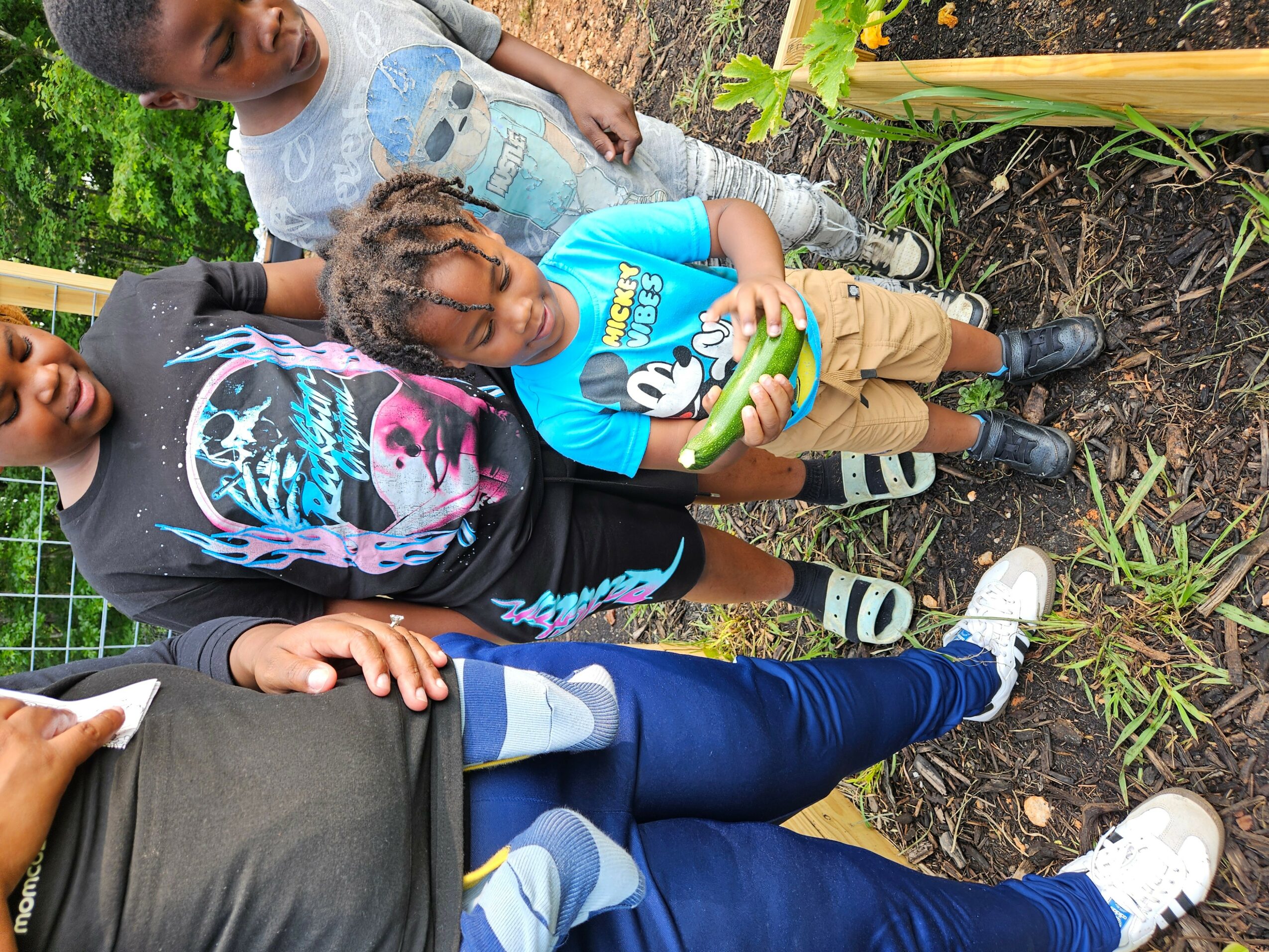 Three children and an adult standing outdoors near plants and soil, with one child holding a green object, all smiling.