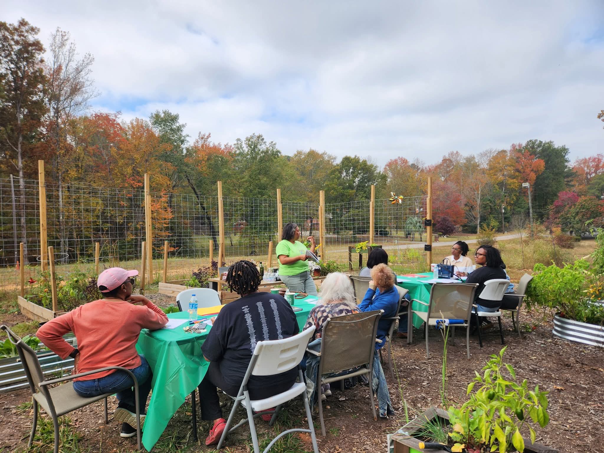 Group of people sitting at tables outdoors, with trees and a fence in the background.