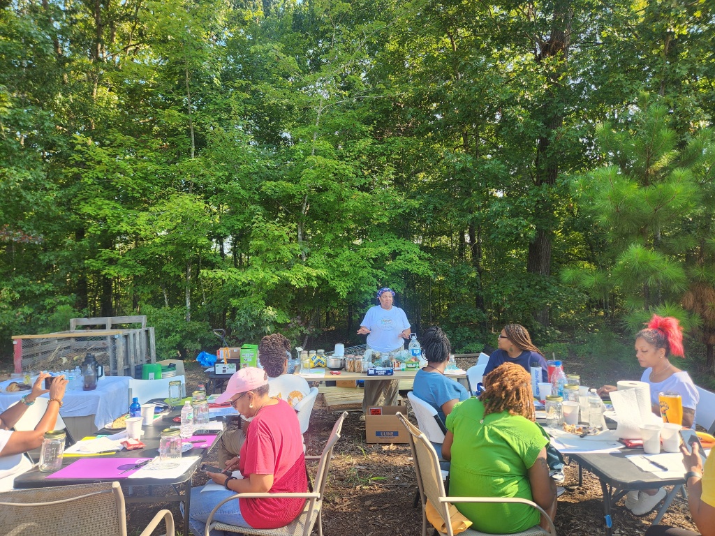 Group of people sitting at tables outdoors in a wooded area, engaging in an activity, with supplies on the tables.