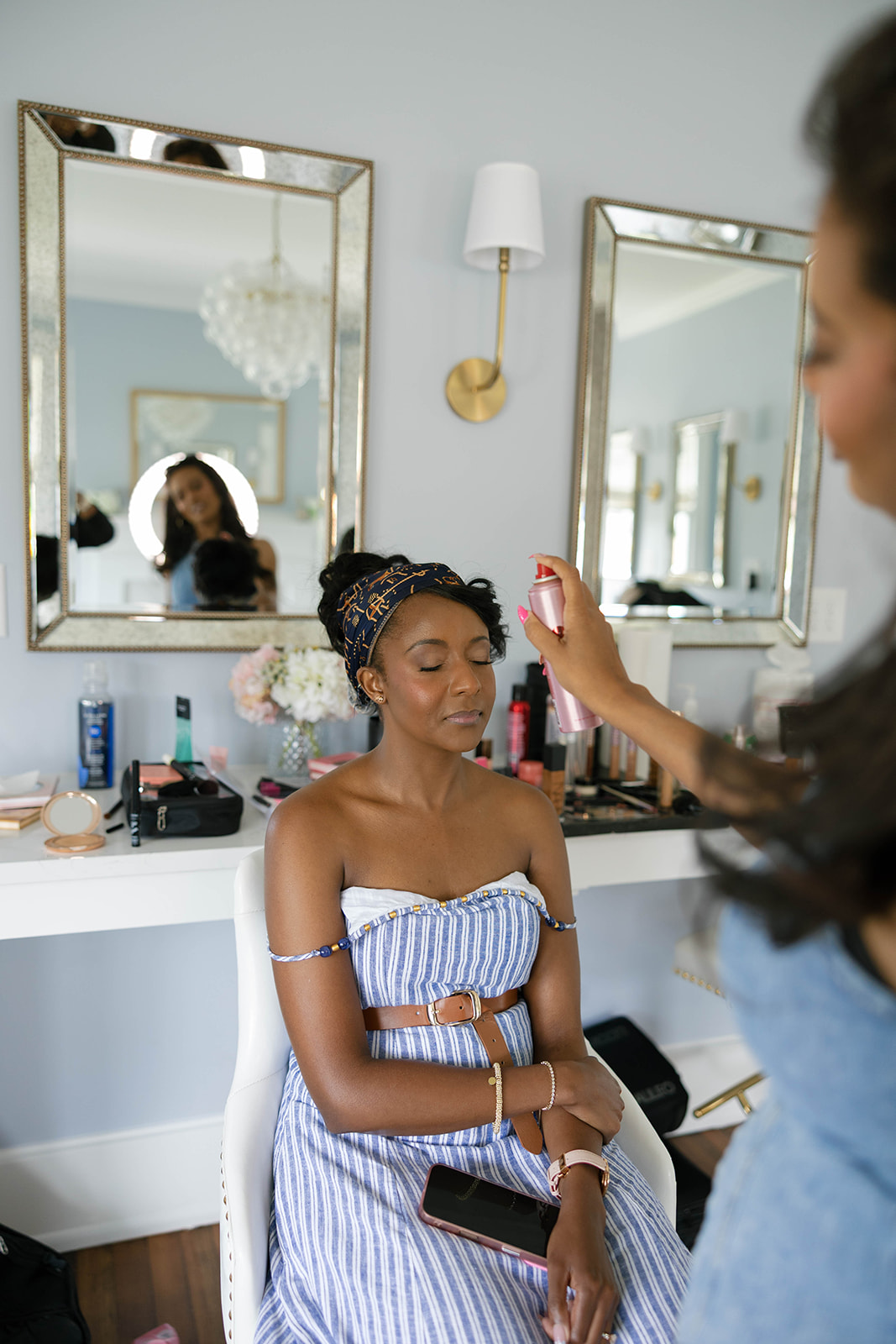 Woman applying makeup to a seated woman in a room with two mirrors, a chandelier, and a wall sconce, with makeup products on the table.