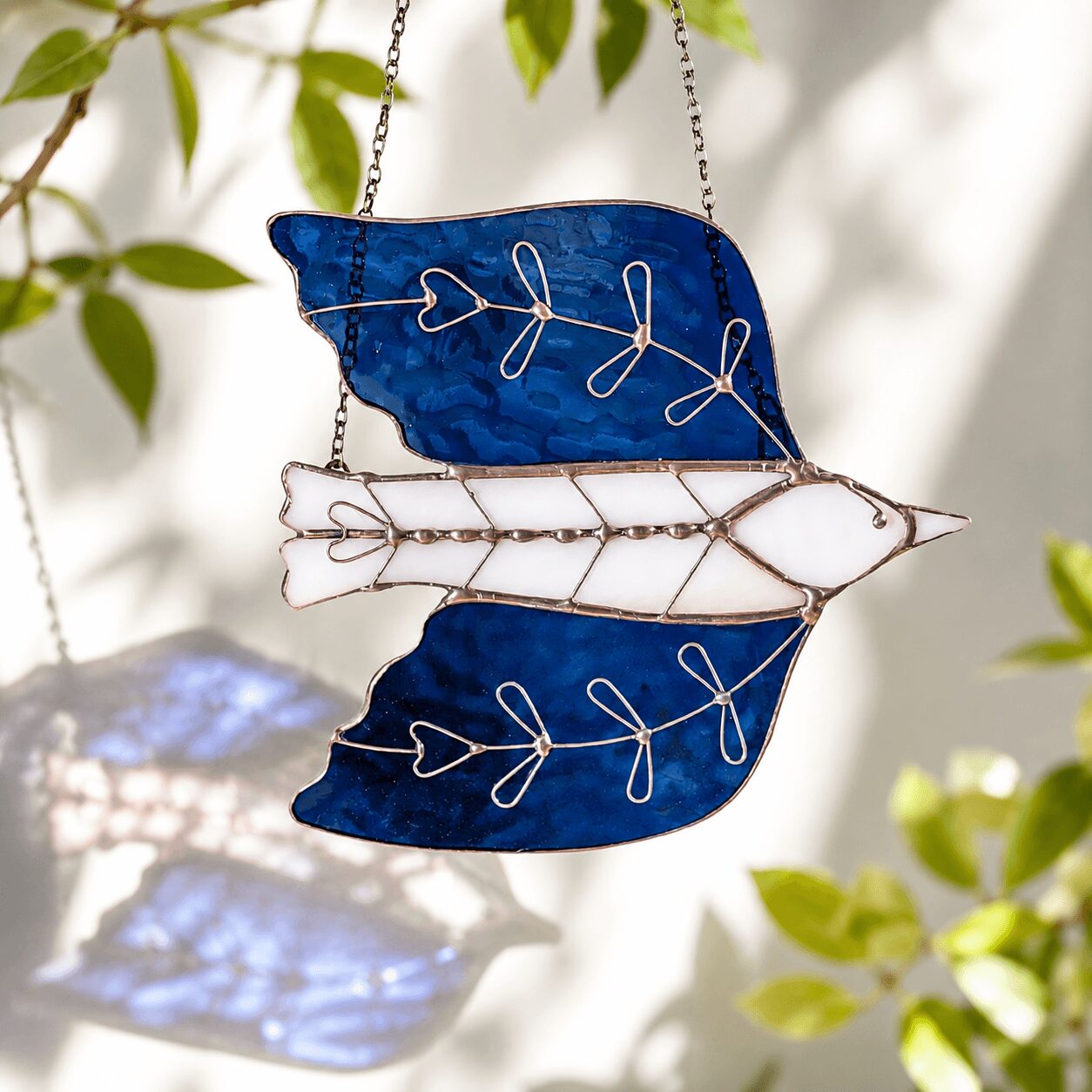 Decorative hanging ornament with three glass pieces resembling leaves and a fish, connected by wire, hanging among green leaves.