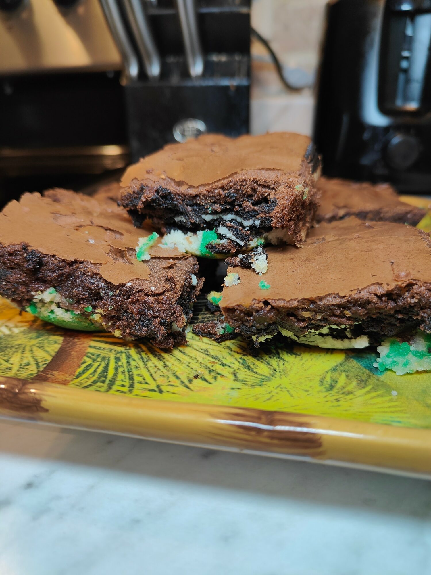 Three chocolate brownies with green and blue sprinkles on a yellow and green patterned plate, kitchen background.
