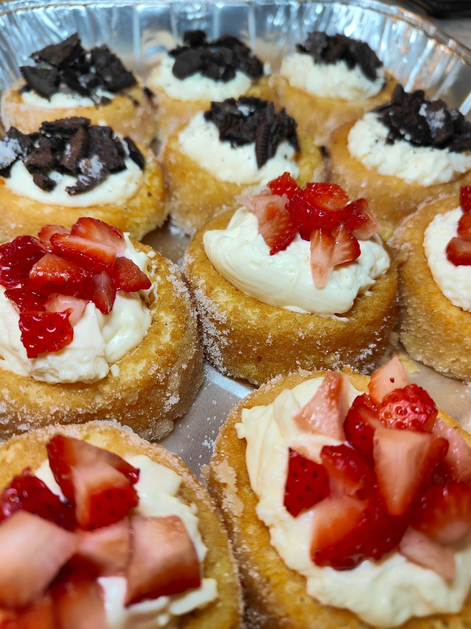 Assorted mini desserts with cream and fruit toppings in a tray, some with chocolate and some with strawberries, on a metallic tray.