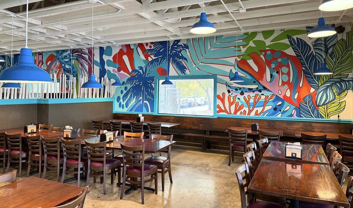 Interior of a restaurant with a colorful tropical mural wall, wooden tables, and chairs, blue pendant lights, and large windows.