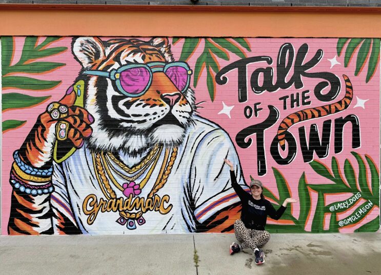 Colorful mural of a tiger wearing sunglasses, jewelry, and a white shirt, with a woman pointing at it, and text reading 'Talk of the Town'.