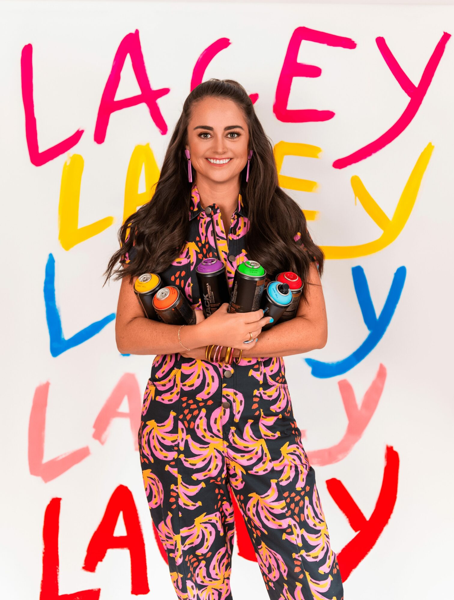 Young woman with long dark hair holding colorful spray paint cans, smiling, with colorful text in the background.