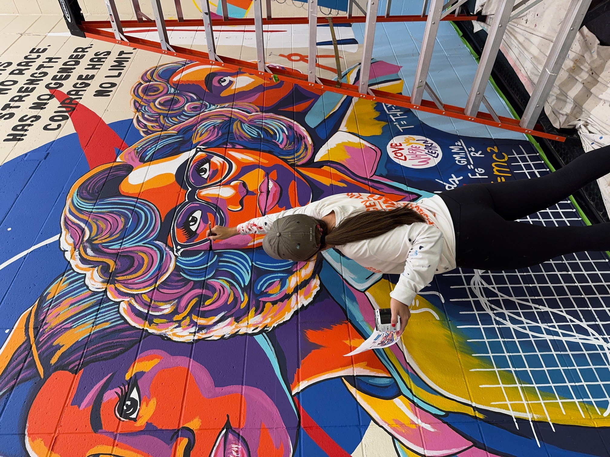 Person painting a colorful mural with abstract and portrait elements on a wall, with text and geometric patterns, indoors.