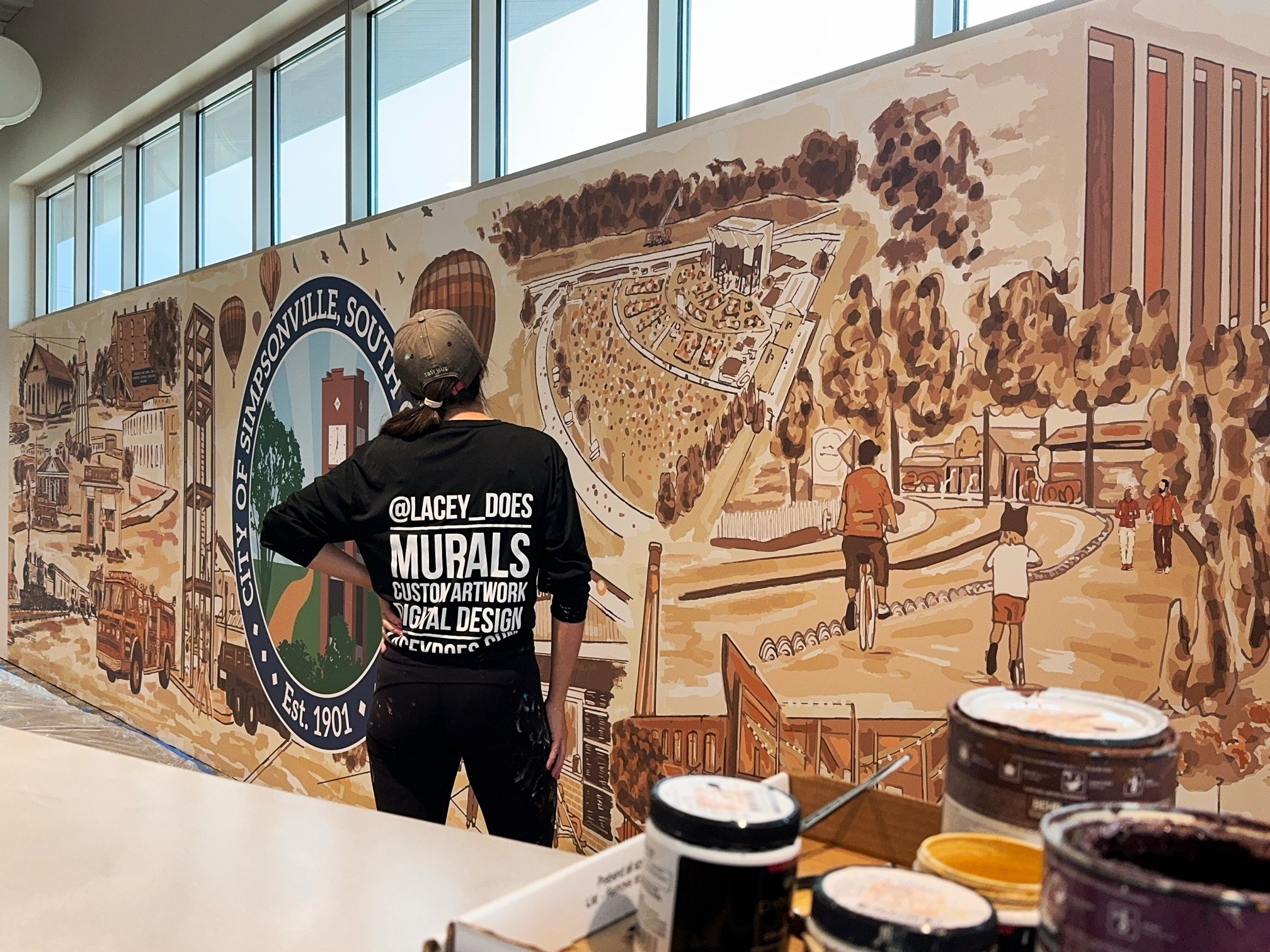 Person viewing a large mural depicting a park, buildings, and people, with a logo on the left side, in a room with large windows.