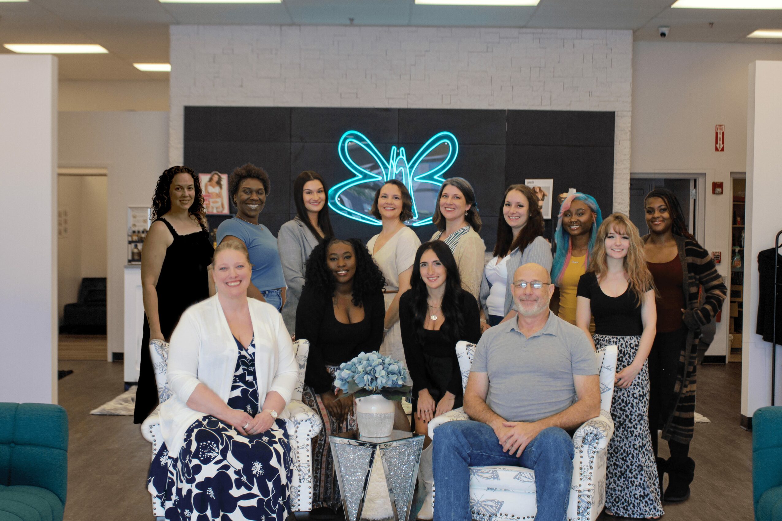 Group of 13 people, mostly women, standing and sitting indoors in front of a black wall with a neon butterfly sign, smiling.