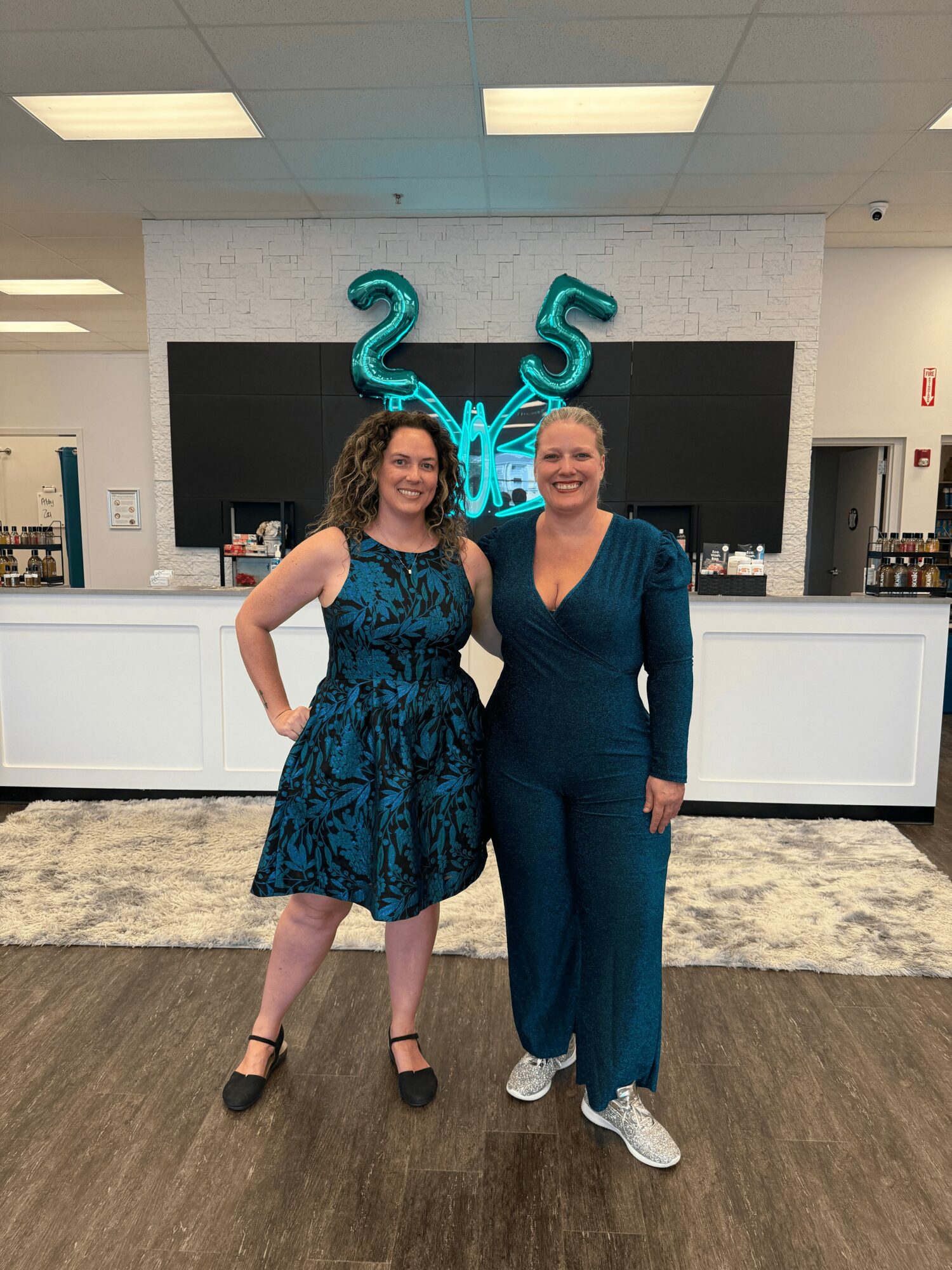 Two women stand in front of a black wall with teal balloon numbers 25 and a teal balloon word, smiling.