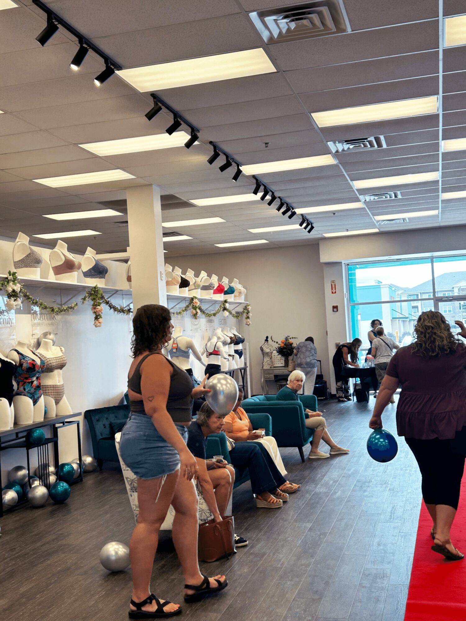 People sitting and standing in a store with mannequins and decorations, some holding balloons, near a large window.