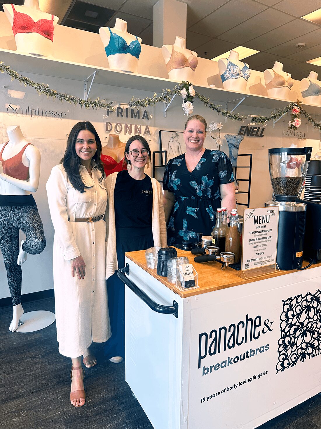 Three women stand behind a counter at a bakery or cafe, smiling, with mannequins and colorful bras displayed above them.