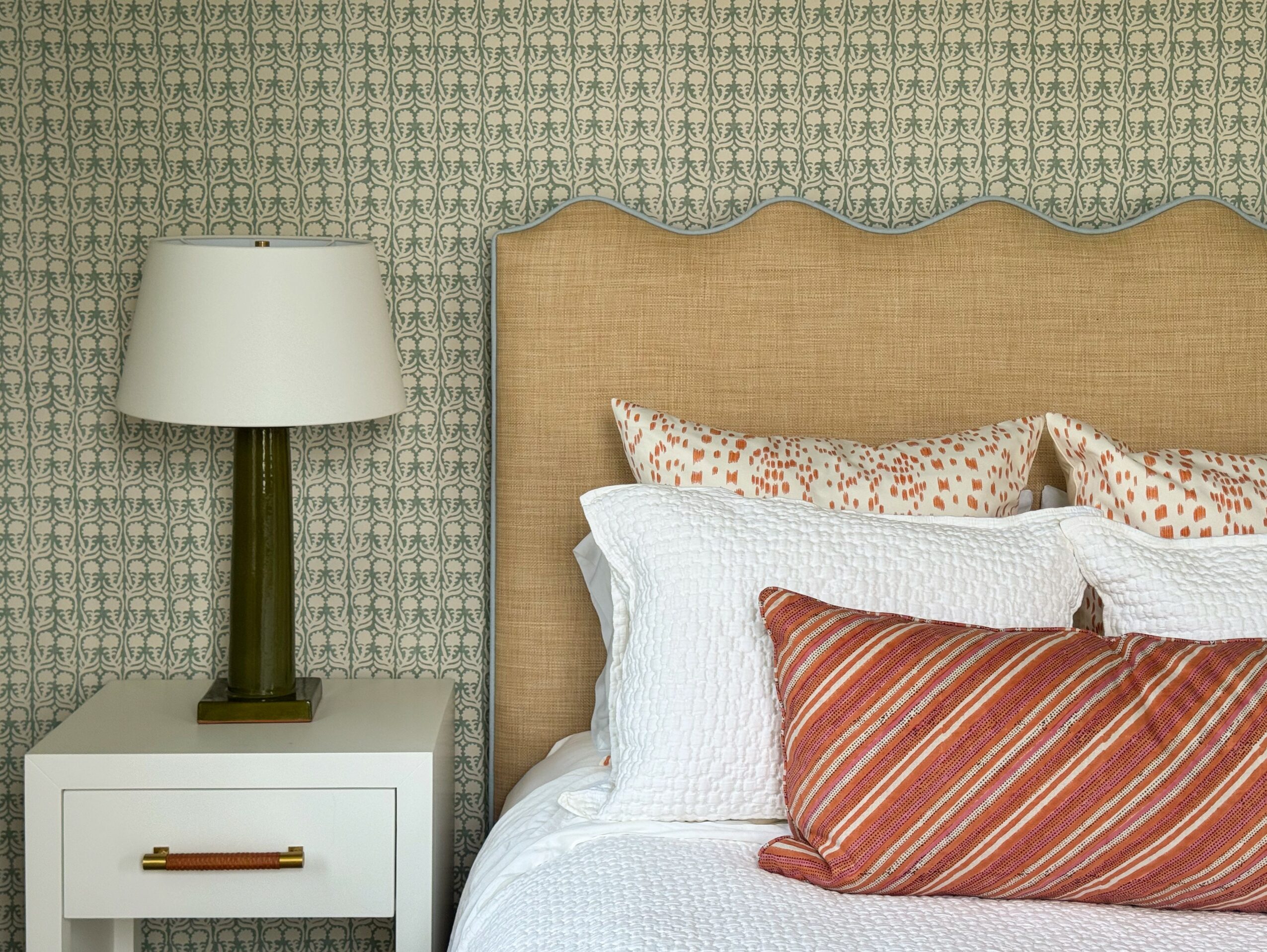 Close-up of a bed with a beige headboard, white and patterned pillows, and a bedside table with a lamp, against patterned wallpaper.