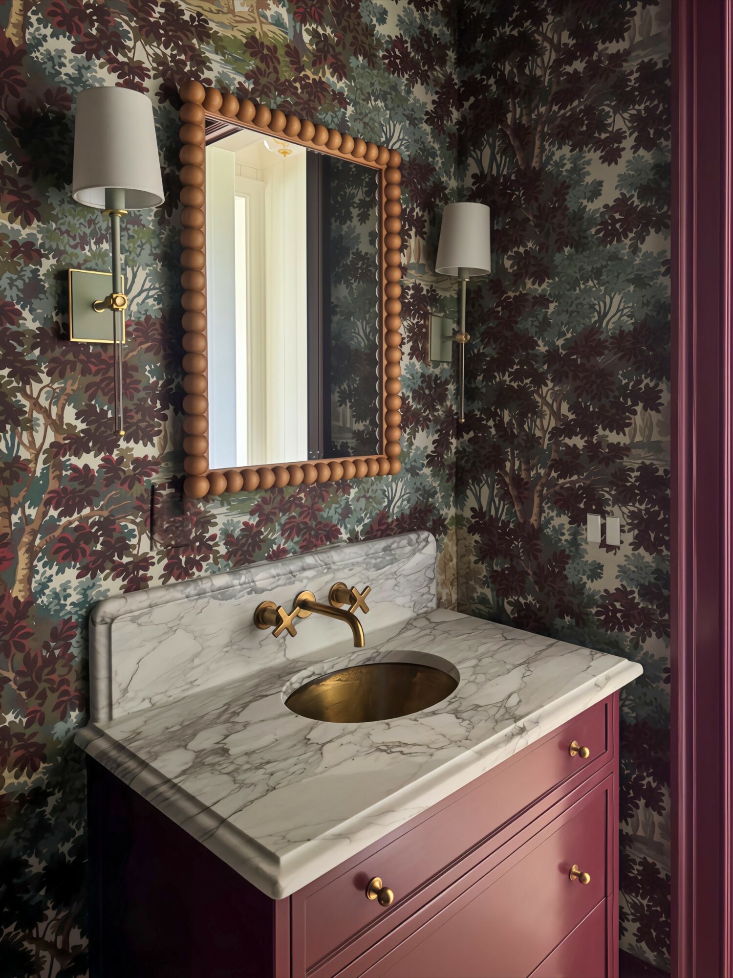 Bathroom vanity with marble countertop, gold faucet, pink cabinet, mirror, and wall-mounted lights, against dark floral wallpaper.