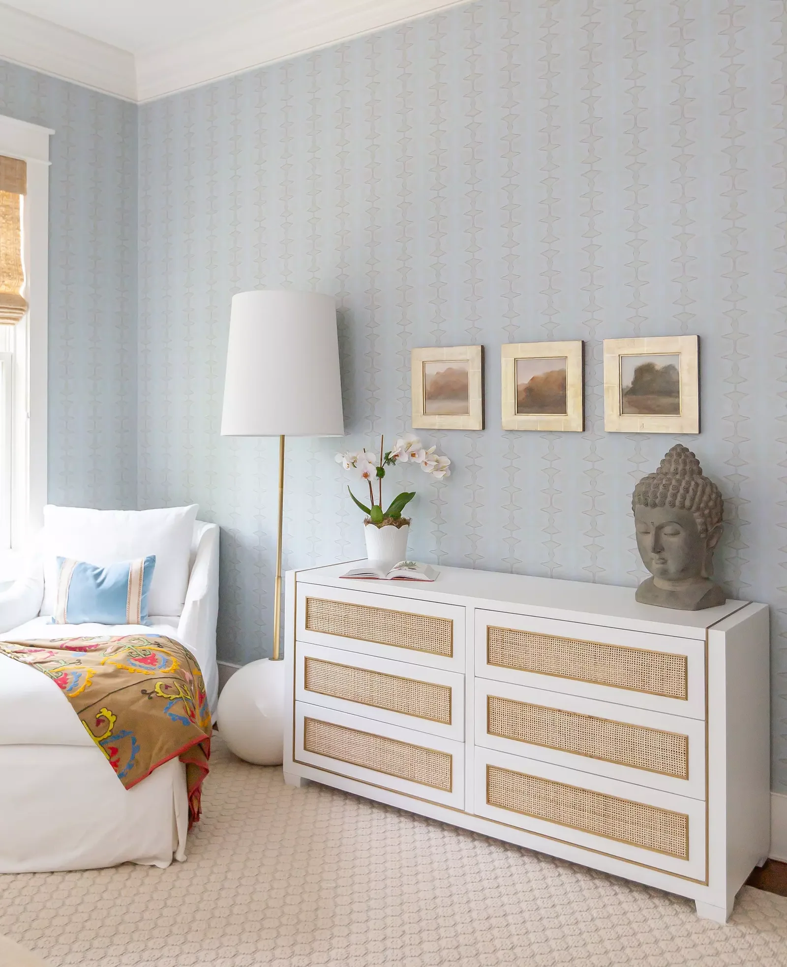Bedroom corner with a bed, white dresser, framed pictures, a lamp, and a decorative bust, with light blue wallpaper.